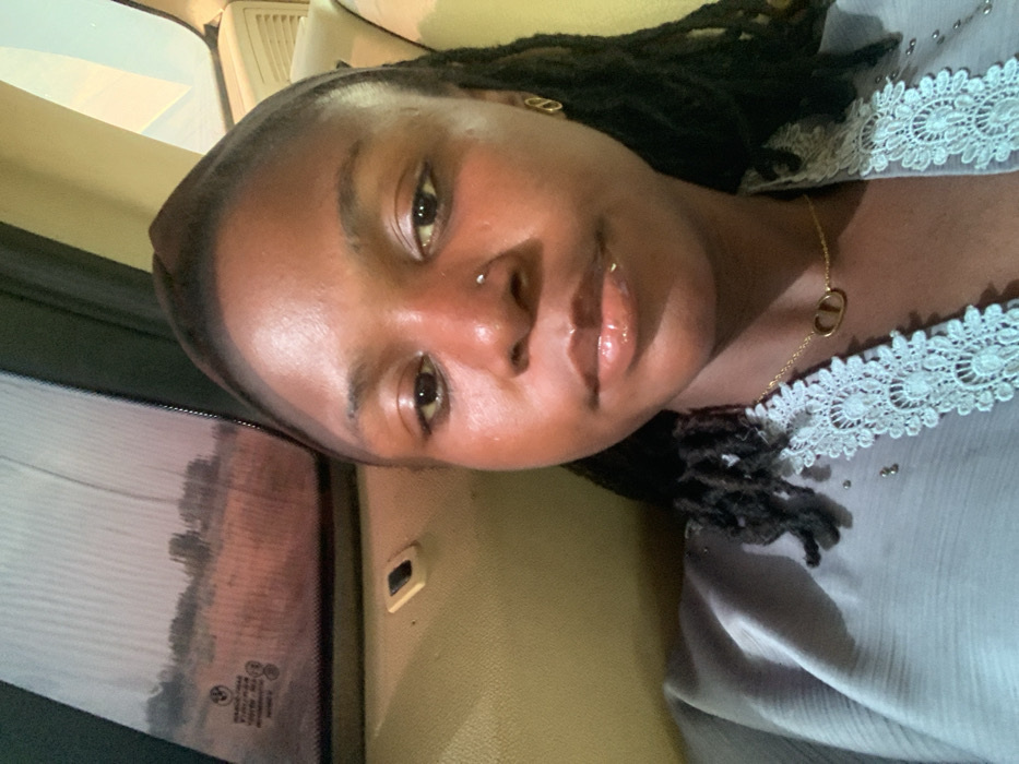 A young adult female is depicted in a posed photograph from within a vehicle, with the stated location being Gadam, Nigeria. She has dark skin, dark dreadlocks, and is wearing a dark headwrap. A small silver stud pierces her right nostril, and her lips are glossy. Her attire includes a light grey garment featuring white lace trim and small metallic studs along the neckline. She wears a thin gold chain necklace with a "CD" pendant and a matching "CD" logo gold earring on her right ear. The vehicle's interior shows a light brown headliner and a side window partially obscured by a textured screen. Through the window, an arid, reddish-brown landscape with sparse, distant vegetation is visible under a diffused sky. A BMW logo is discernible on the window glass. The individual looks directly forward.