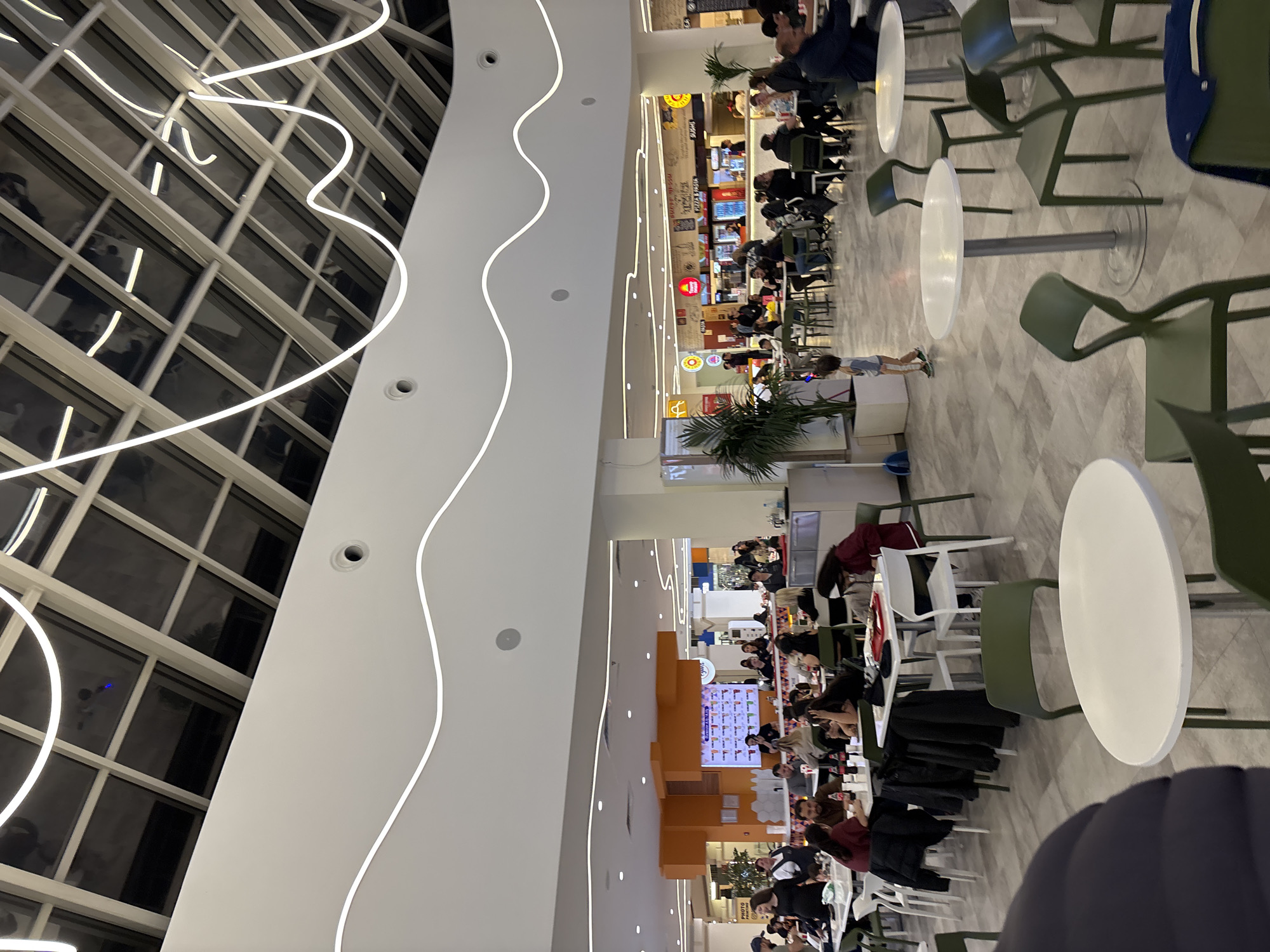This indoor shot depicts a food court in a shopping mall in Yerevan, Armenia. The scene is well-lit with natural light coming through the large, gridded windows on the left. The ceiling is high and white, featuring wavy, illuminated lines.
Numerous tables and chairs, mostly light green, are arranged for dining. People are seated at some tables, eating and socializing. There's a variety of food vendors visible, with signage in Armenian or Russian. The environment appears busy and active. There is a young child running near the tables. There are also plants in the area.