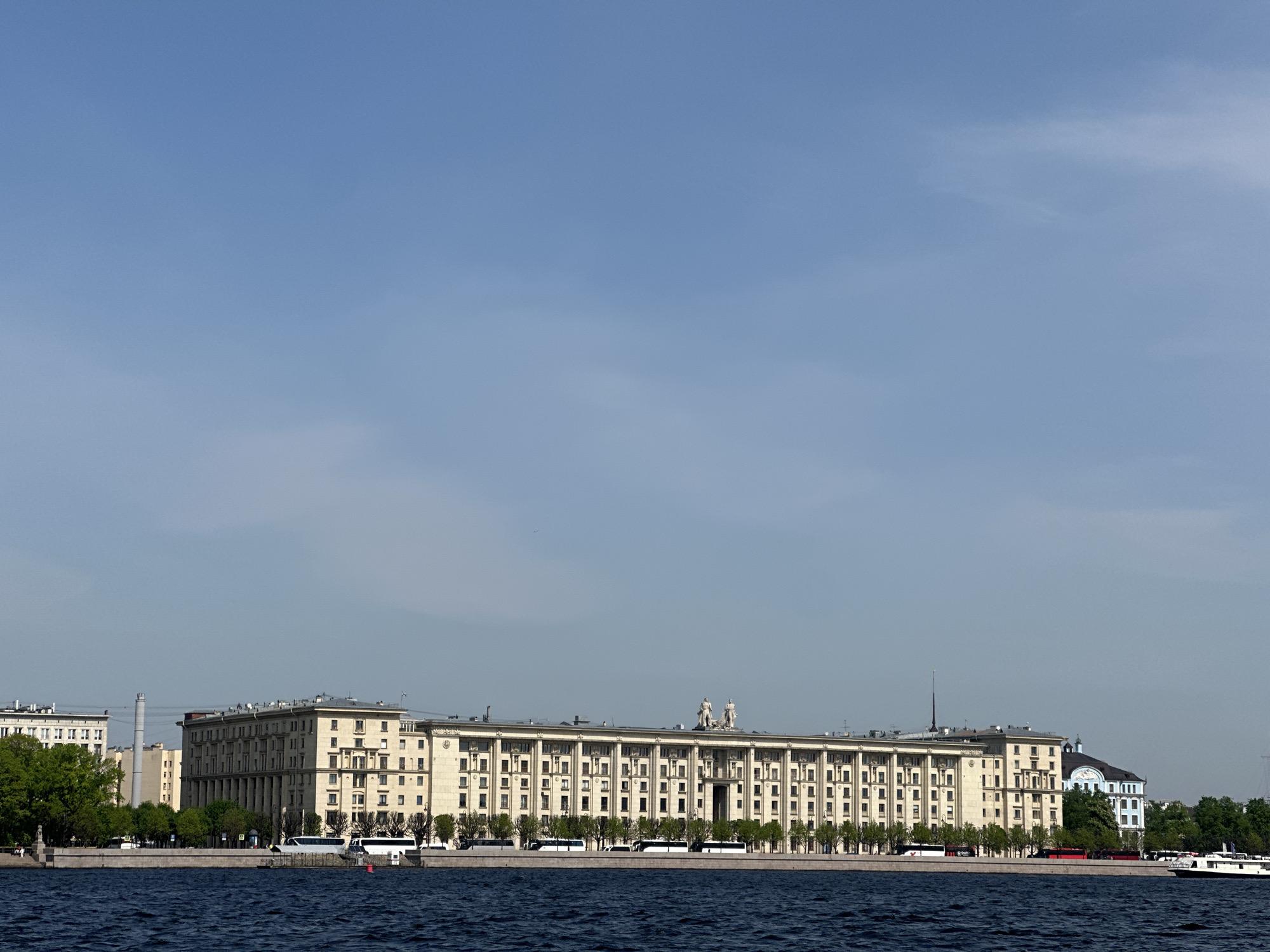 The image shows a long, beige-colored building with classical architectural elements such as columns and statues on its roof, located in Saint Petersburg, Russia. The building is situated along a waterfront with a stone embankment. In front of the building, several white buses are parked. A few boats are visible on the water. Trees are planted along the embankment. The sky is light blue with some scattered clouds. In the background, other buildings can be seen, including one with a blue and white facade. The water in the foreground is dark blue.