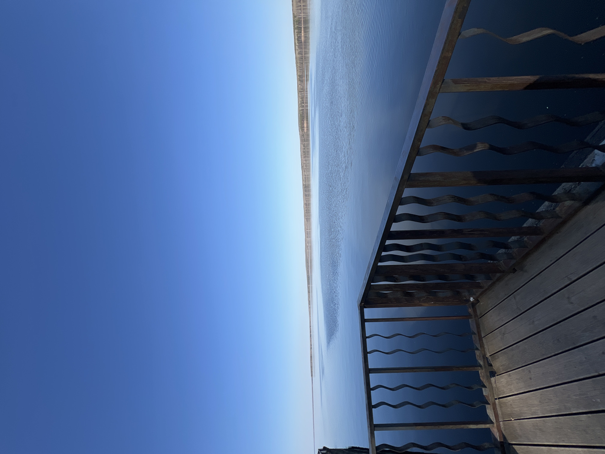 This image, taken from a raised platform, features a body of water reflecting the sky. The water stretches across the frame with a thin strip of land visible on the far side. A clear, blue sky dominates the left side of the image. A wooden railing, with a wavy metal insert, is visible on the right side, as well as the wooden deck. The image may be from Chaikovsky, Russia, given the request of location.