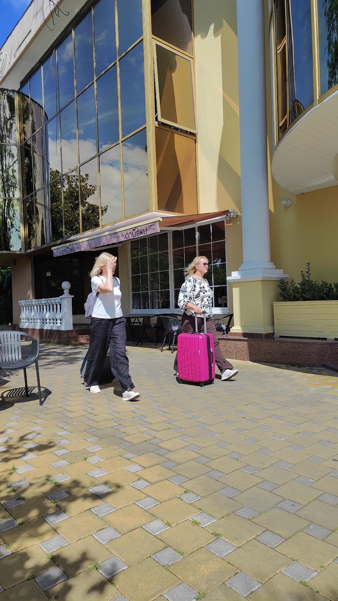 Two women are walking on a paved walkway in front of a building with a glass facade. The woman on the left is wearing a white t-shirt and black pants and is pulling a dark rolling suitcase. The woman on the right is wearing a black and white patterned shirt and brown pants and is pulling a bright pink rolling suitcase. A white pillar stands to the right of the women. The building has awnings with Cyrillic lettering, suggesting it is a cafe or shop. The walkway is made of light yellow and gray rectangular paving stones. Trees are visible in the reflection of the building's windows, and shadows from trees are cast on the walkway. The scene appears to be in Sochi, Russia, possibly near a hotel or resort given the architectural style and signage.