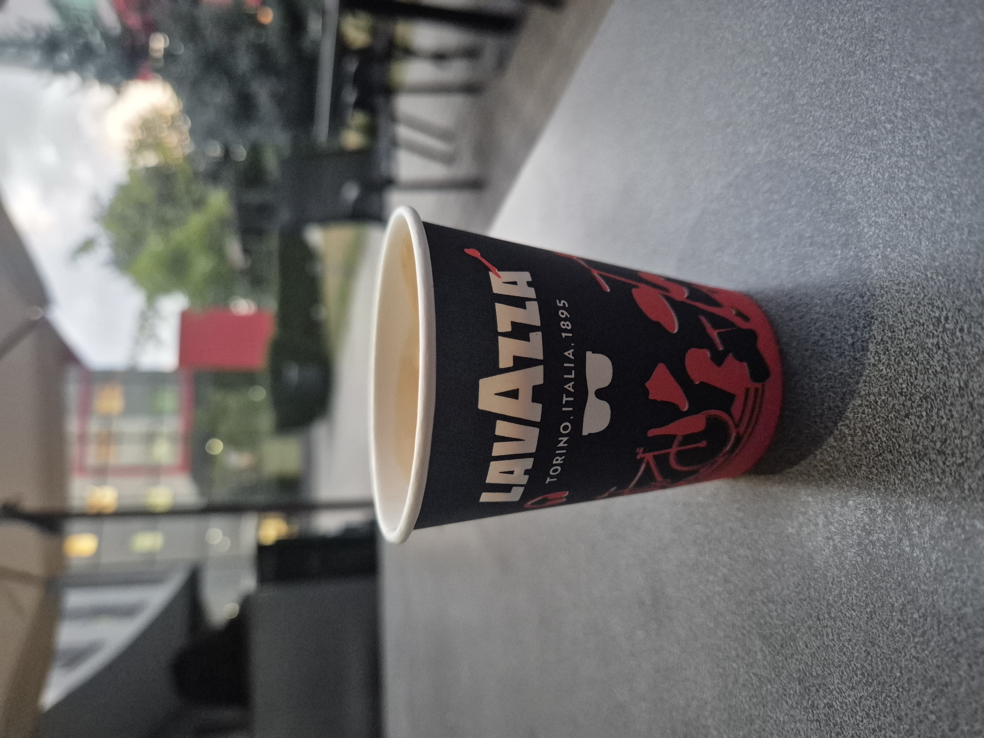 A Lavazza coffee cup is positioned on a textured gray surface, with its logo and design visible. The cup contains a light brown beverage. In the background, out of focus, are buildings, trees, and outdoor seating areas, suggesting an urban or commercial outdoor setting. The lighting indicates it is daytime, with possibly overcast conditions.