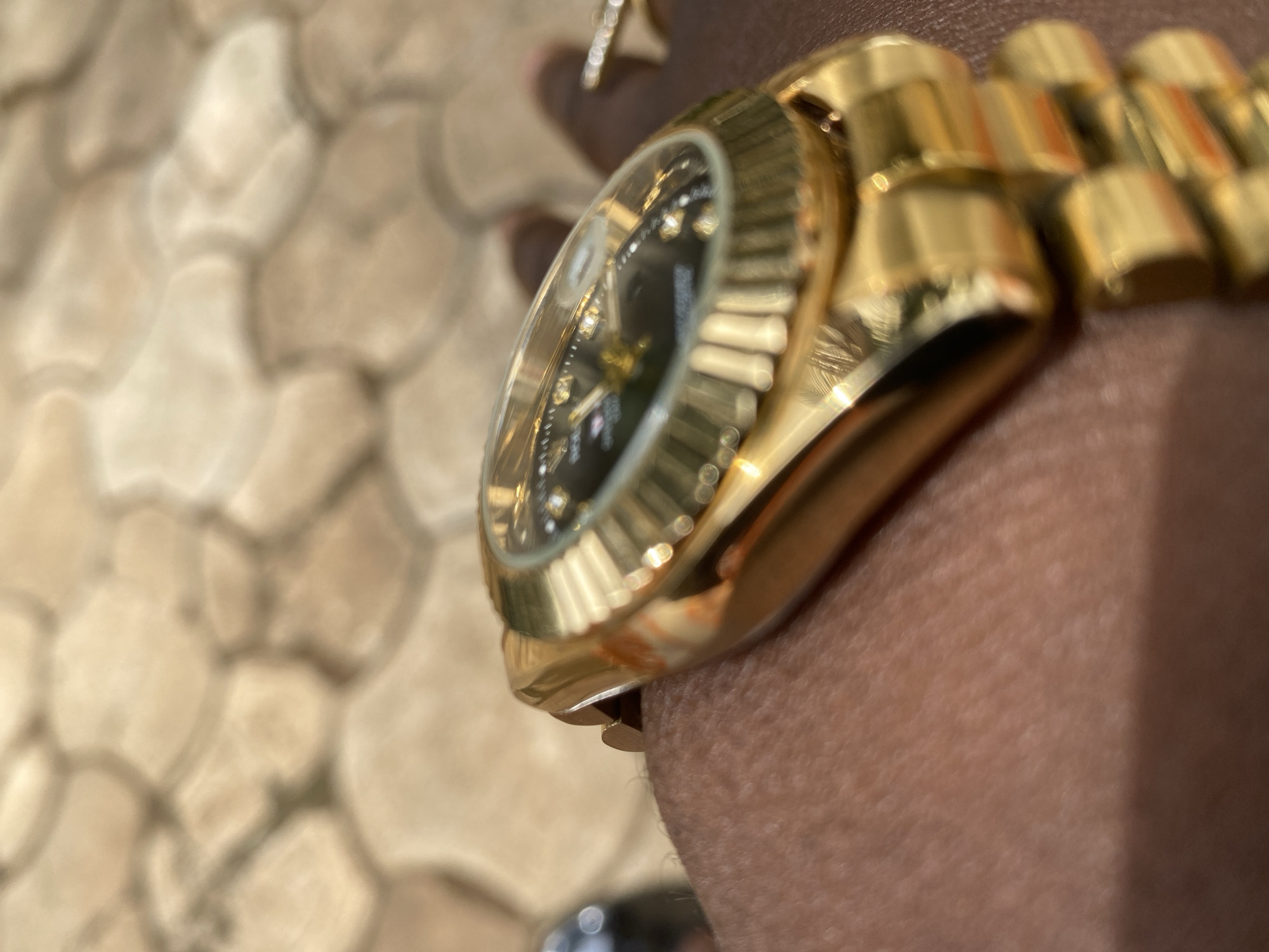 A close-up shot depicts a gold watch with a black face resting on dark skin. The watch has a fluted bezel and a gold band. A ring is visible on the finger. The background is a blurred image of textured, beige tiles. The city of Maigizo, Nigeria is not discernible from the image.