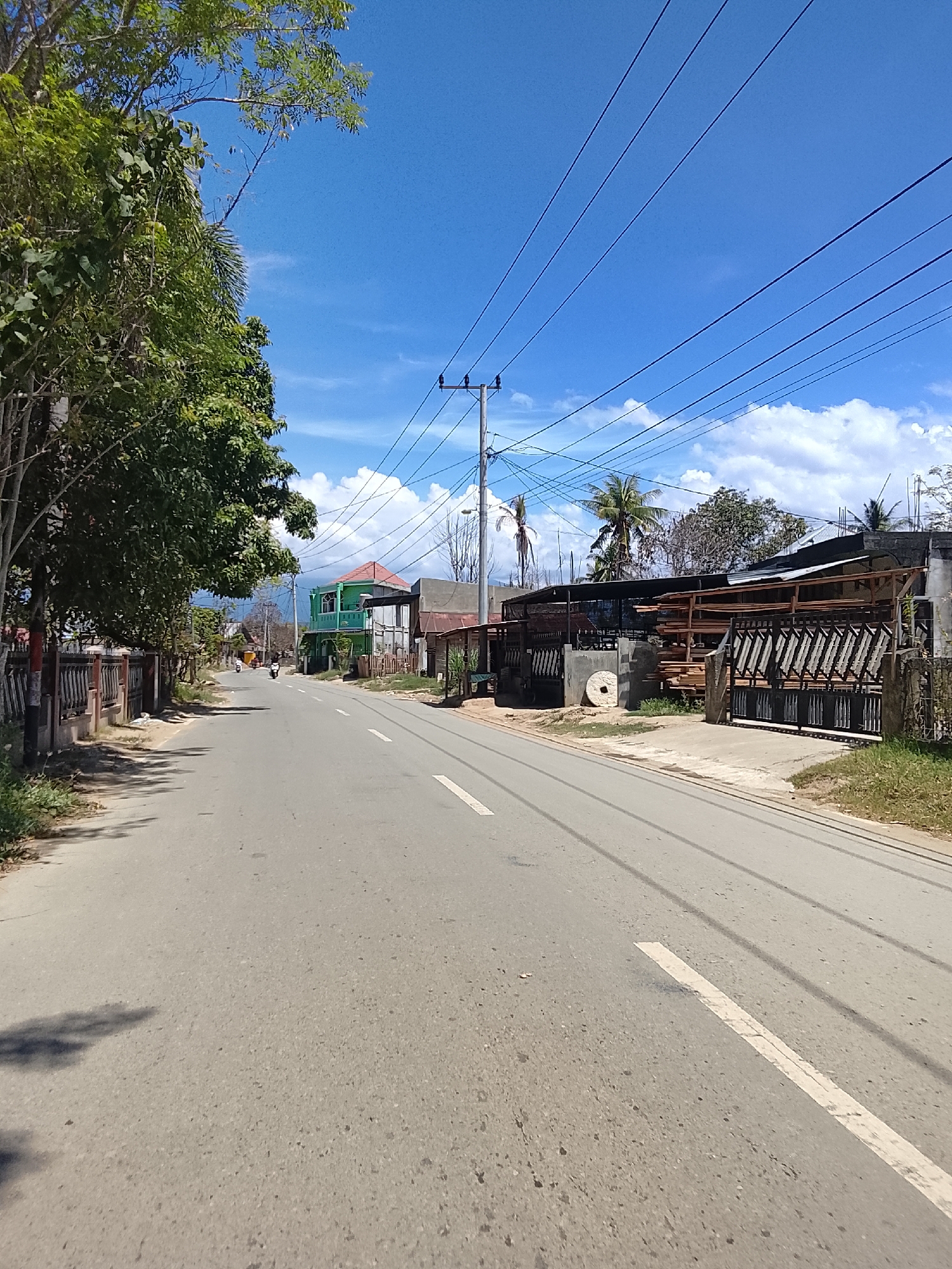 The image depicts a street scene in Blang Cut, Indonesia, under a bright blue sky with scattered white clouds. The road, paved with asphalt and marked with a white dashed line, stretches into the distance, curving slightly to the right. Lush green trees line the left side of the road, casting dappled shadows on the pavement. On the right side of the road, a low fence is visible, followed by a building with a corrugated metal roof and a large, dark metal gate. Beyond this, a two-story green building with a red roof stands prominently. Electrical wires are strung across the sky, supported by utility poles. In the background, the landscape appears somewhat rural, with additional buildings and vegetation visible. Two motorcycles are seen on the road, suggesting ongoing activity. The overall impression is that of a typical sunny day in a developing urban or suburban area in Indonesia.