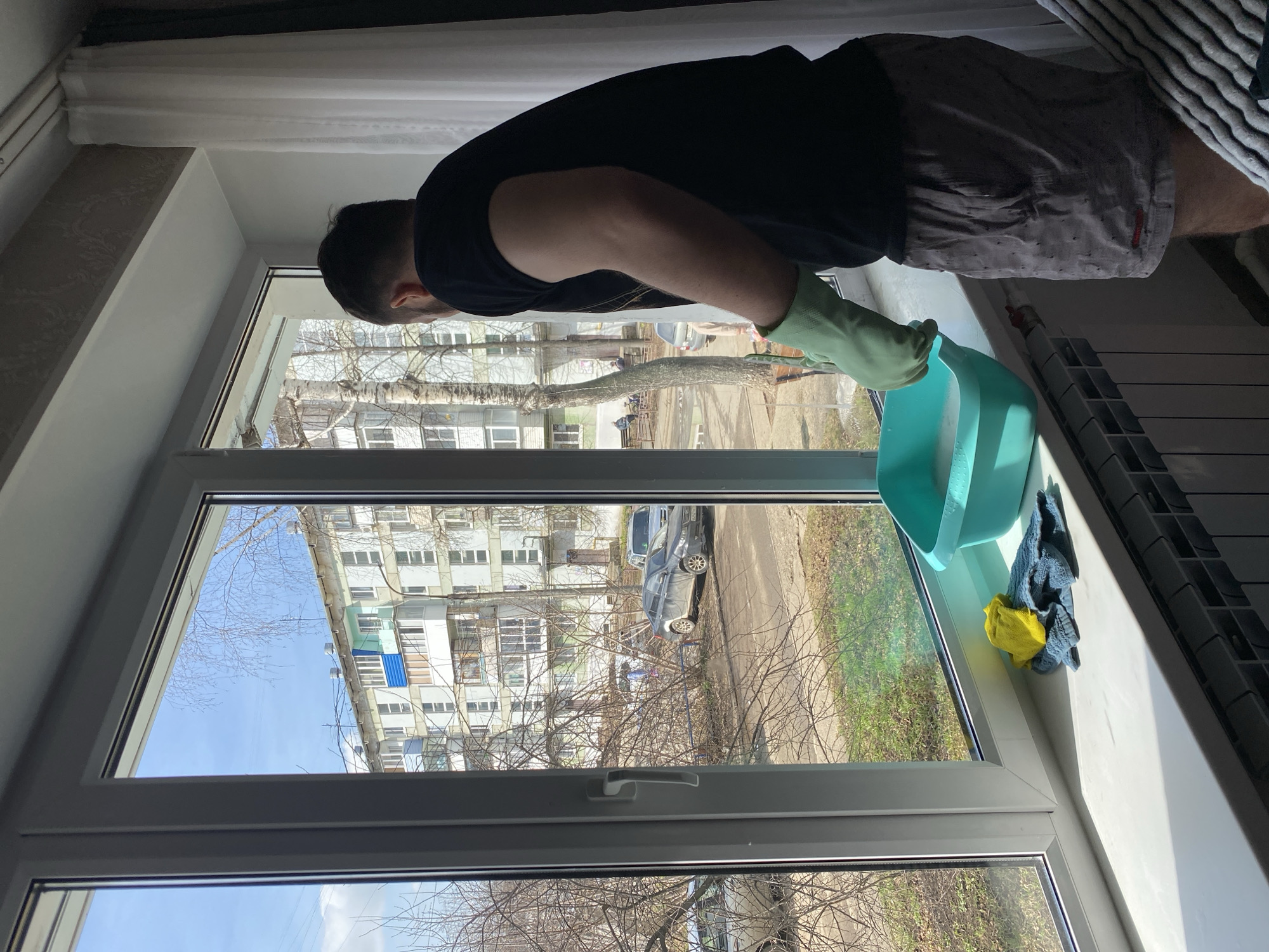 A person in a black t-shirt and patterned shorts is cleaning a window in Chaikovsky, Russia. They are wearing a green glove on one hand and holding a light blue bucket with cleaning solution. A yellow cloth and a blue towel are on the windowsill. Through the window, a multi-story building, trees without leaves, a car, and a blue sky can be seen.