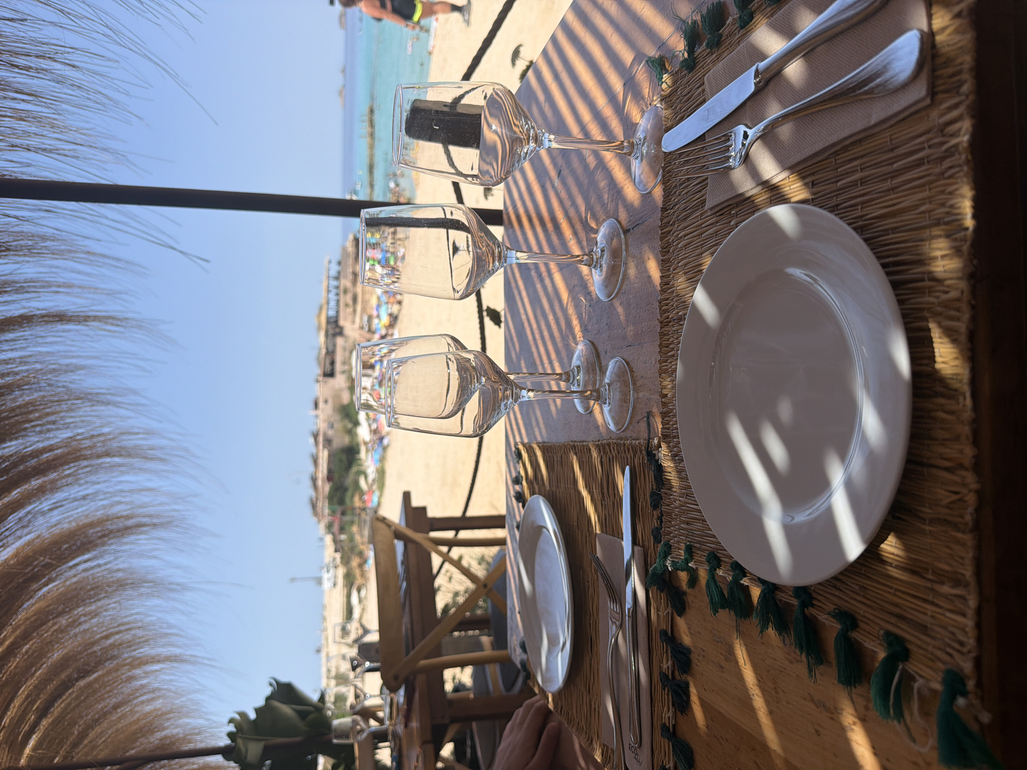 This image depicts a beachfront restaurant setting in Sant Josep de sa Talaia, Spain. The scene is set on a wooden table covered with straw placemats adorned with green tassels. On each placemat, a white plate is set, with cutlery (a fork and a knife) to the right. Four empty wine glasses are arranged on the table. In the background, the turquoise waters of the Mediterranean Sea are visible, along with a sandy beach populated by people and beach umbrellas. The sky is clear and blue. The restaurant appears to be open-air, with a thatched roof overhead casting striped shadows on the table. A person's hand is visible on the left side of the frame, suggesting someone is seated at the table.