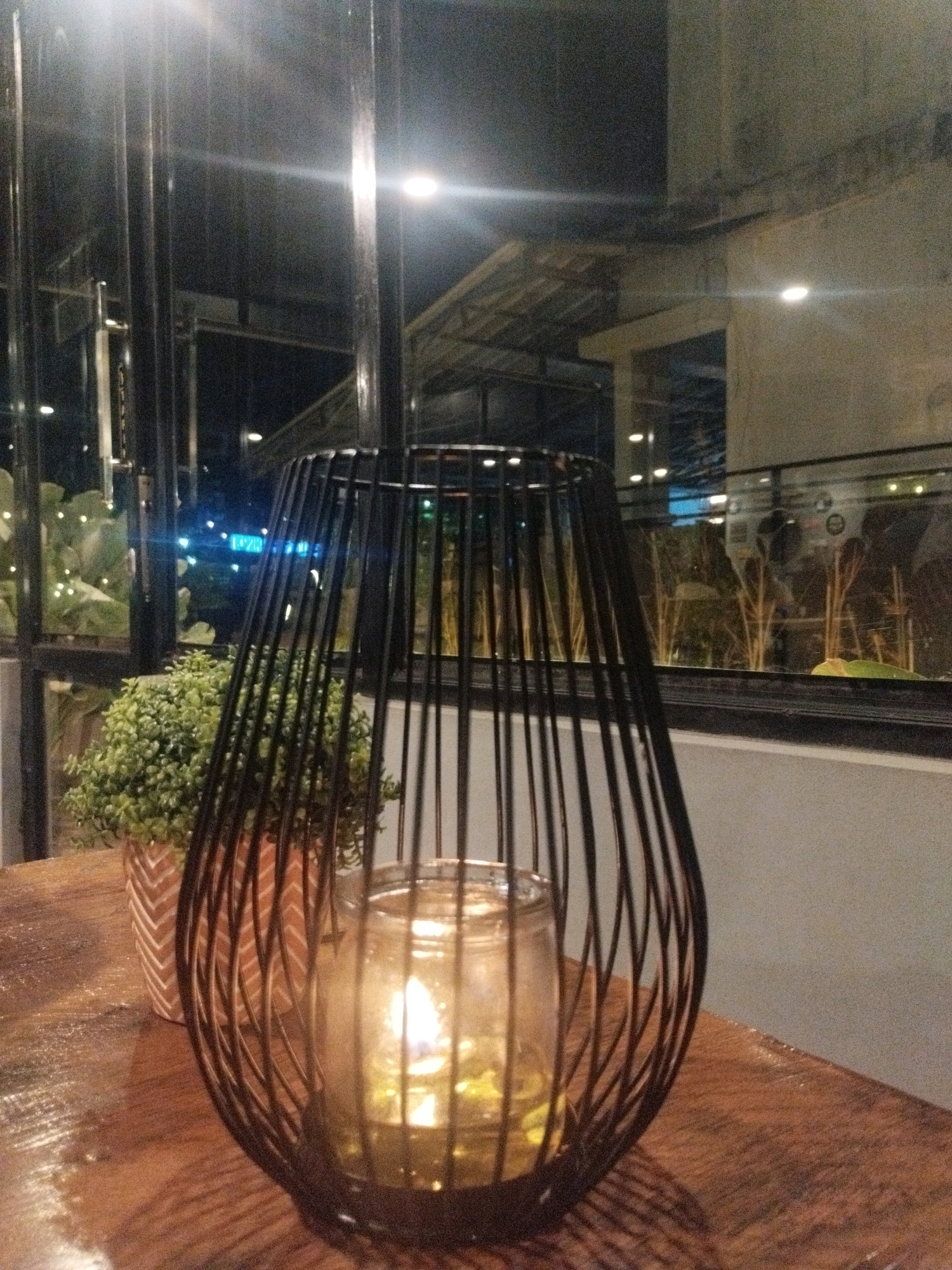 The image captures a dimly lit indoor scene, likely a cafe or restaurant, with a focus on a decorative black metal lantern holding a lit candle. The lantern is placed on a wooden table in the foreground, which shows some texture and possibly light speckles. To the left of the lantern is a small potted plant with lush green foliage, in a patterned terracotta-colored pot.
Through a large glass window behind the table, a nighttime cityscape is visible. The exterior features include architectural structures with visible lighting, some appearing to be part of a building complex. In the background, there are tall, thin plants or reeds, creating a naturalistic element to the outdoor space. The lighting in the scene is primarily from the candle within the lantern and artificial lights from the exterior, creating a warm and atmospheric ambiance. The reflection on the glass suggests the presence of more interior lights and possibly people.
The location is identified as Sungailiat, Indonesia, which implies the scene is set within this city. The overall impression is one of a cozy, inviting establishment, perhaps offering a relaxing dining or social experience in Sungailiat. There are no people directly visible in the frame, focusing the attention on the objects and the interplay of light and shadow.