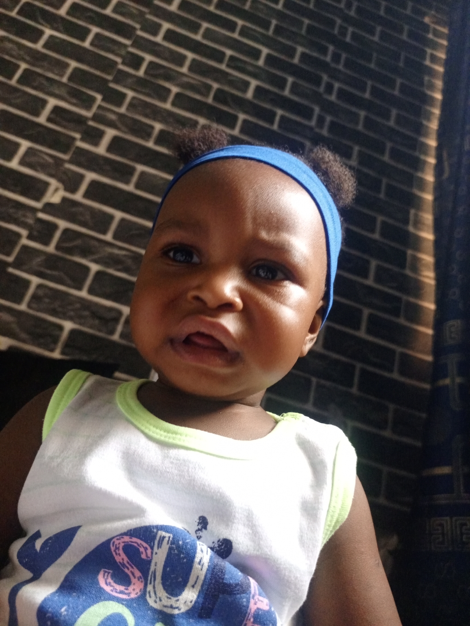 A close-up, chest-up portrait shows a baby indoors in Keffi, Nigeria. The baby, with brown skin and chubby cheeks, looks directly towards the camera with a somewhat serious or slightly pouting expression; their lips are slightly parted, revealing a hint of the tongue. Dark eyes are open wide.
The baby wears a bright blue fabric headband, under which small, dark, curly hair bunches are visible on top of their head. They are dressed in a white sleeveless top featuring a light green trim around the neck and armholes. A colorful graphic design is present on the front of the shirt, with the partial letters "SU" clearly visible in shades of blue and pink.
The background behind the baby consists of a dark grey or black wall with a prominent brick pattern. To the far right of the frame, a dark, patterned fabric, likely a curtain or drape, can be seen, partially in shadow. The lighting appears to be from an indoor source, casting some subtle shadows and highlighting the baby's face. The overall impression is a candid shot of a curious or slightly bewildered infant.