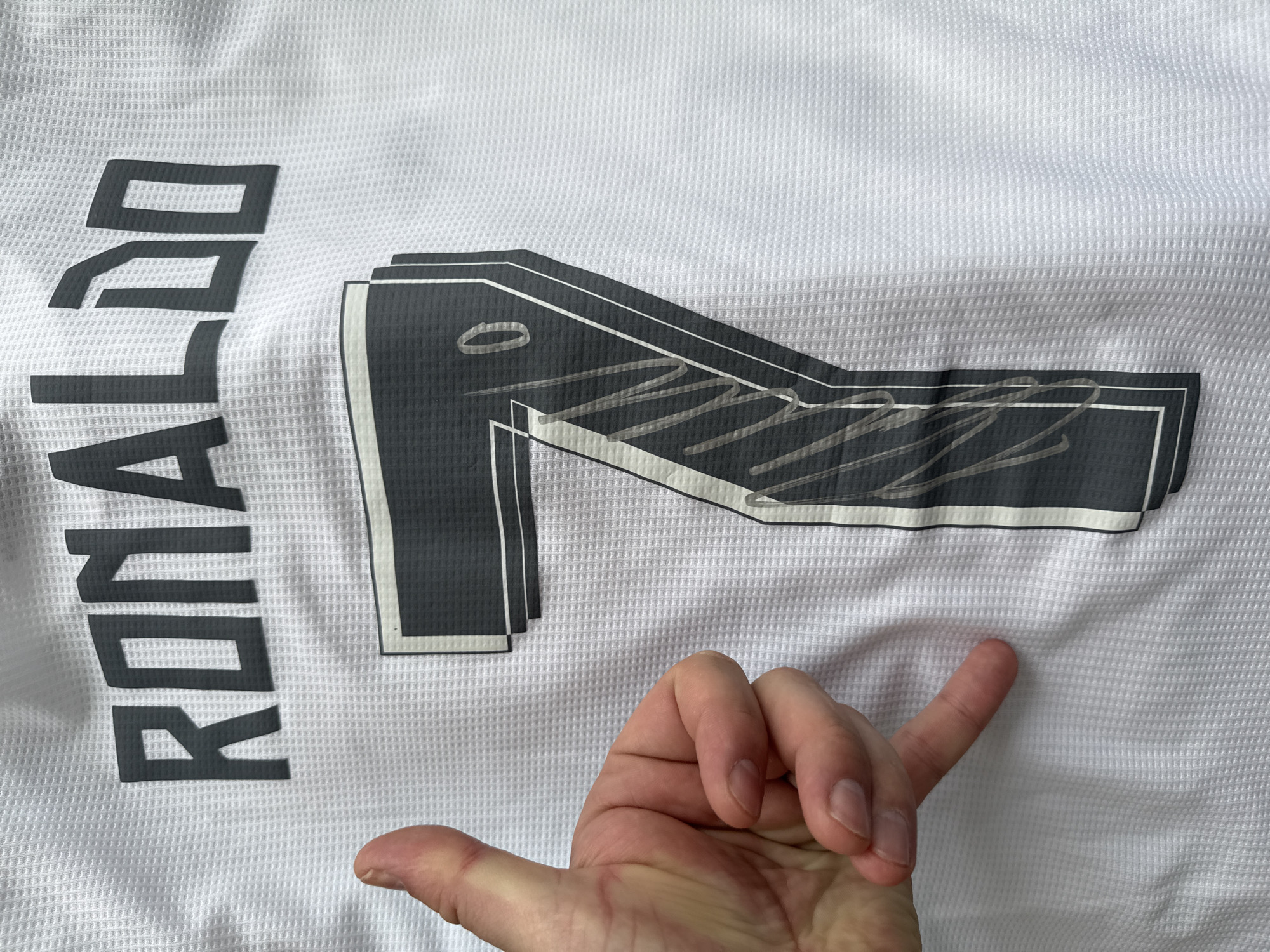The image shows a white soccer jersey with the name "RONALDO" printed vertically in black on the left side. Next to the name, a large number "7" is displayed, also in black, with a white border. The number appears to have a textured or patterned fill within its outline. In the lower-right corner of the image, a hand points toward the fabric of the jersey. The background consists of the white, textured surface of the jersey, which appears to be made of a breathable, athletic material. The scene's location is not discernible, and no information suggests it is in Vladivostok, Russia.