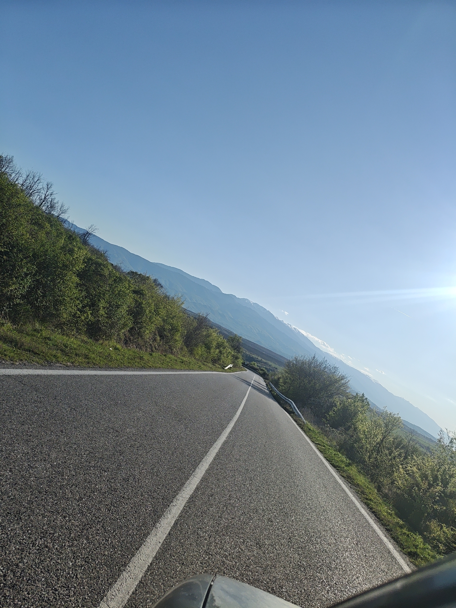 A tilted, medium-shot photograph depicts a road winding alongside a mountain range under a clear, blue sky.
The road is made of asphalt, with a solid white line dividing the lanes. Vegetation is present on both sides of the road, consisting of green grass and trees. In the background, a mountain range is visible, with the sunlight appearing to the right of the image. Parts of a car (such as the hood and side) are visible at the bottom of the photo, suggesting that the picture was taken from inside of the car.