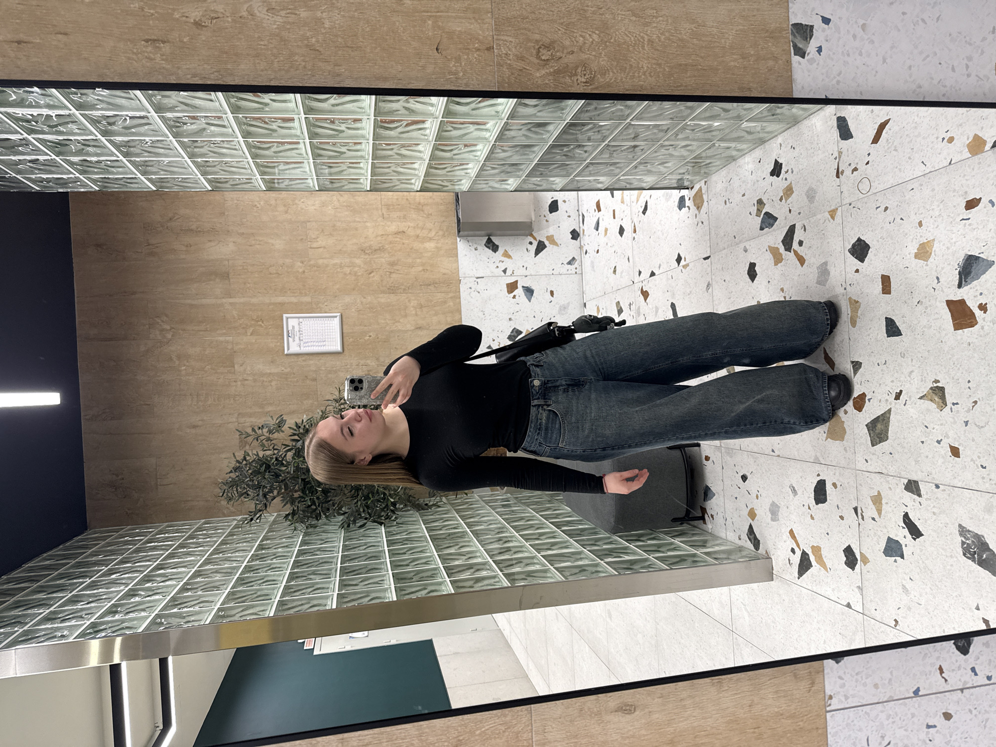 A young female is captured in a full-length mirror selfie within an indoor commercial or public space in Sochi, Russia. She stands centrally, facing left with her head slightly tilted, holding a light-colored smartphone in her raised right hand to photograph her reflection. She has long, straight, light brown hair and is dressed in a black long-sleeved top, dark blue wide-leg jeans, and dark, closed-toe footwear. A black cross-body bag hangs from her left shoulder. The flooring consists of white terrazzo with irregular chips in shades of black, grey, brown, and blue. The background features a light brown wood-textured wall on the right, and a translucent wall constructed from glass blocks immediately to its left. Behind the subject, to the left of the glass block wall, a dark grey upholstered bench and an artificial plant with dark green leaves are visible. Reflected in the large mirror is a dark teal wall, another section of the wood-textured wall, and linear ceiling lighting fixtures. A white framed document is mounted on the reflected wood-textured wall, above a metallic waste receptacle.