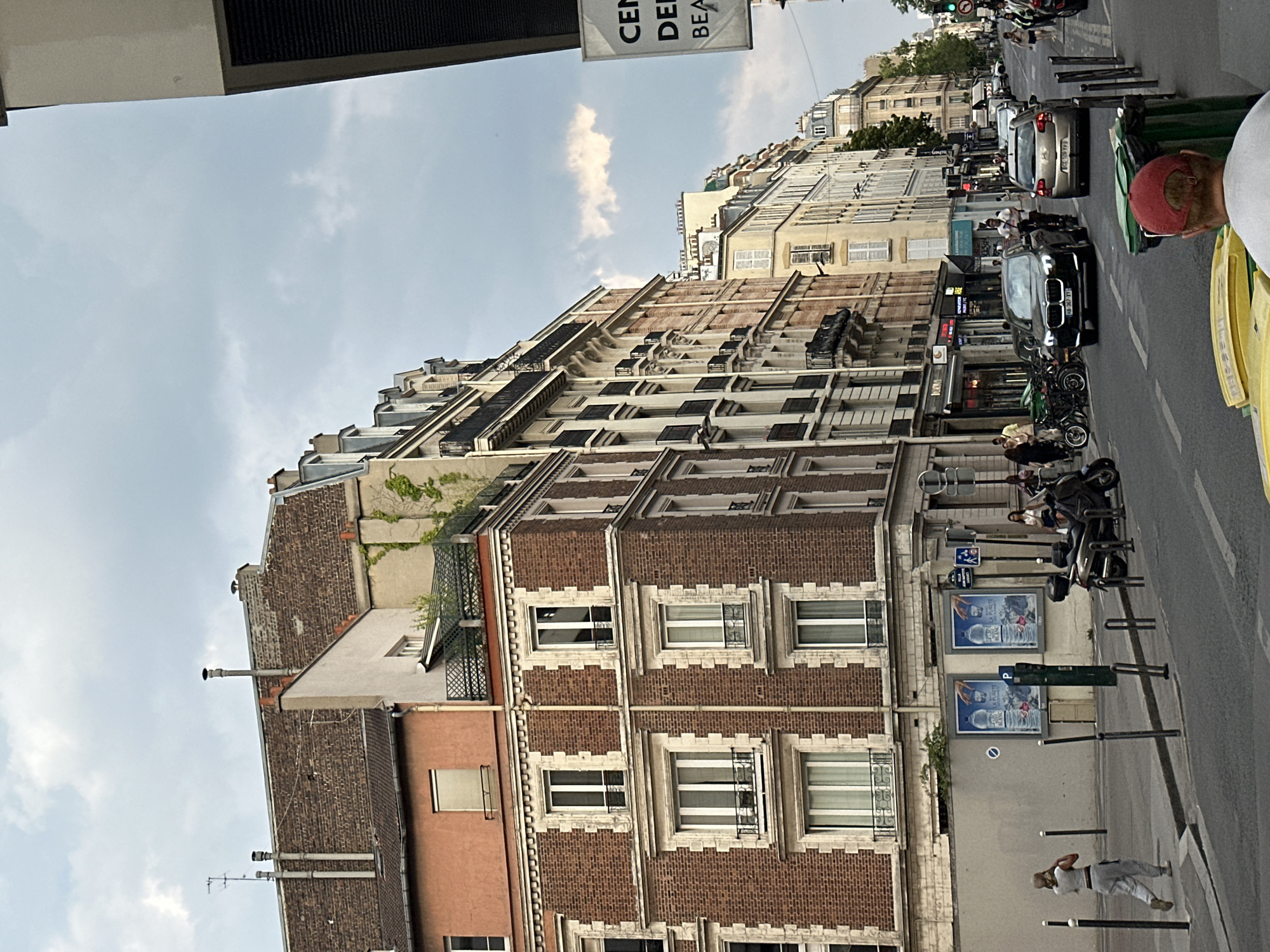 This is a street-level, eye-level shot taken in Paris, France, on a cloudy day. The perspective looks down a street lined with multi-story buildings. The buildings have traditional Parisian architecture, with stone facades, mansard roofs, and many windows. Some buildings are brick, others are cream-colored stone.
In the foreground, on the right side, a person with a red cap is partially visible, facing away from the camera. They are looking at large green and yellow trash bins. The street is paved with asphalt, with a sidewalk on the left. There are metal bollards along the sidewalk.
On the left side of the street, a woman is walking away from the camera, dressed in light-colored casual clothing. Behind her, there are two large posters on a grey wall, depicting bottles of water. A green utility box is also present.
The street is moderately busy with traffic, including cars and motorcycles. Several vehicles are visible, some parked and some in motion. In the distance, the street continues, lined with more buildings and trees. Traffic lights and street signs are also visible. The overall impression is of a typical urban scene in Paris, capturing the everyday life of the city.