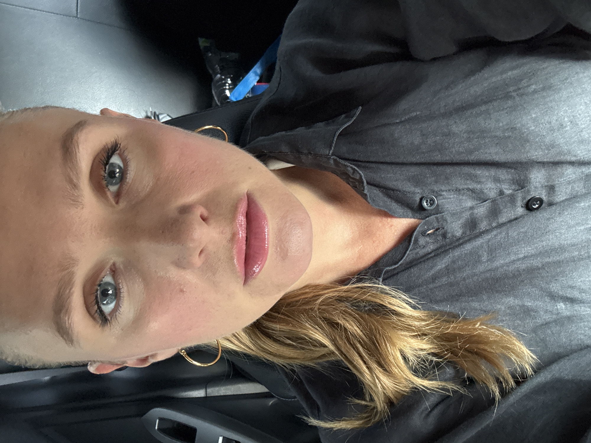 A close-up, angled portrait features a woman with blonde to light brown, wavy hair and light blue-grey eyes, looking directly at the camera. Her complexion is fair, and her lips are glossy with a pink tint. She is wearing a dark grey or charcoal button-up shirt, which is slightly unbuttoned at the top. A single gold hoop earring is visible on her left ear. Her hair falls over her shoulder and chest, resting on the fabric of her shirt. The background is dark and out of focus, appearing to be the interior of a car, with subtle hints of dark surfaces and possibly a seatbelt or car panel. Her expression is neutral and direct. There is no visible text in the image.