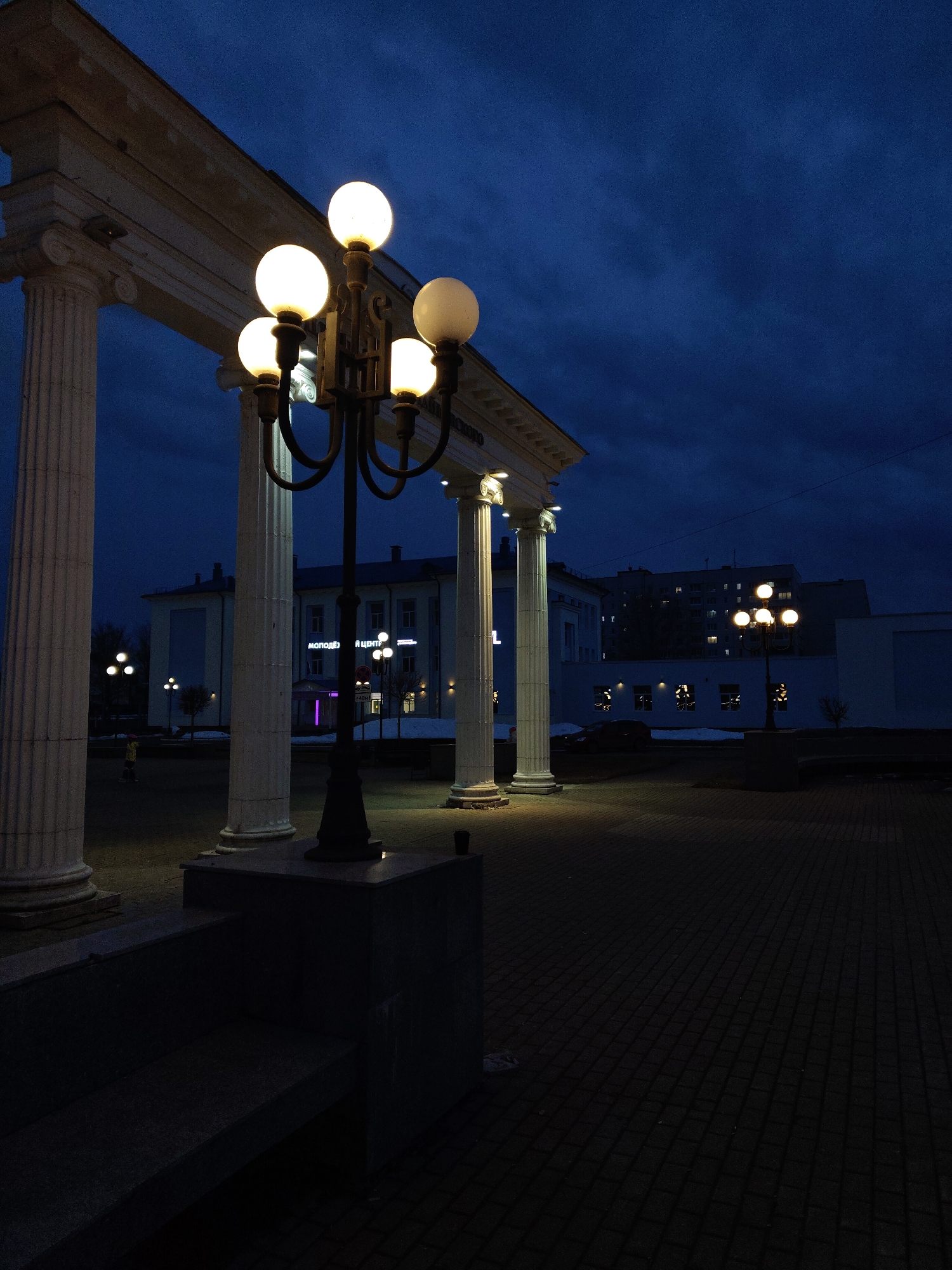 Captured at night, the image showcases a cityscape of Unknown, Unknown. The frame is dominated by a colonnade-like structure composed of white pillars topped with a horizontal entablature, suggesting classical architectural influence. A vintage-style lamppost with multiple globe-shaped lights stands prominently in the foreground, positioned on a raised platform or base. In the background, buildings of varying heights are visible, some illuminated by internal or street lighting. The sky above is dark, hinting at dusk or nightfall, with cloud cover contributing to the overall dimness of the scene. The ground is paved with what appears to be brick or stone tiles, adding to the urban environment. Another similar lamppost can be seen further in the background, contributing to the ambient lighting of the area. The perspective suggests the photograph was taken from a stationary position at ground level, emphasizing the depth and scale of the urban landscape.