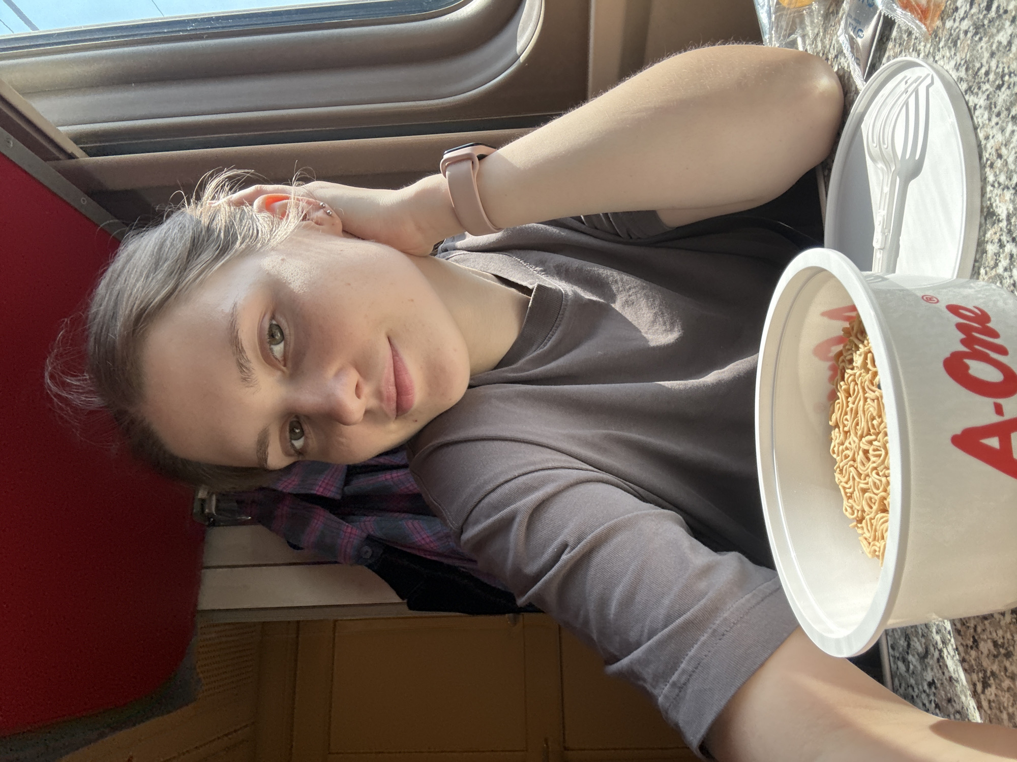 A young Caucasian woman with light brown hair pulled back is seated on a train, likely in Chudovo, Russia, judging by the context of the location. She is looking at the camera with a slight smile and has her left hand resting on her head. She is wearing a plain grey t-shirt and a light pink fitness tracker on her right wrist. In front of her, on a speckled surface that appears to be a table, is a white cup of instant noodles. A white plastic fork and plate are also visible. The background shows the interior of a train carriage, with wooden paneling, a red upholstered ceiling, and a window with brown trim. A plaid shirt is hanging on a hook behind her. The lighting suggests it is daytime, with sunlight streaming in from the window.