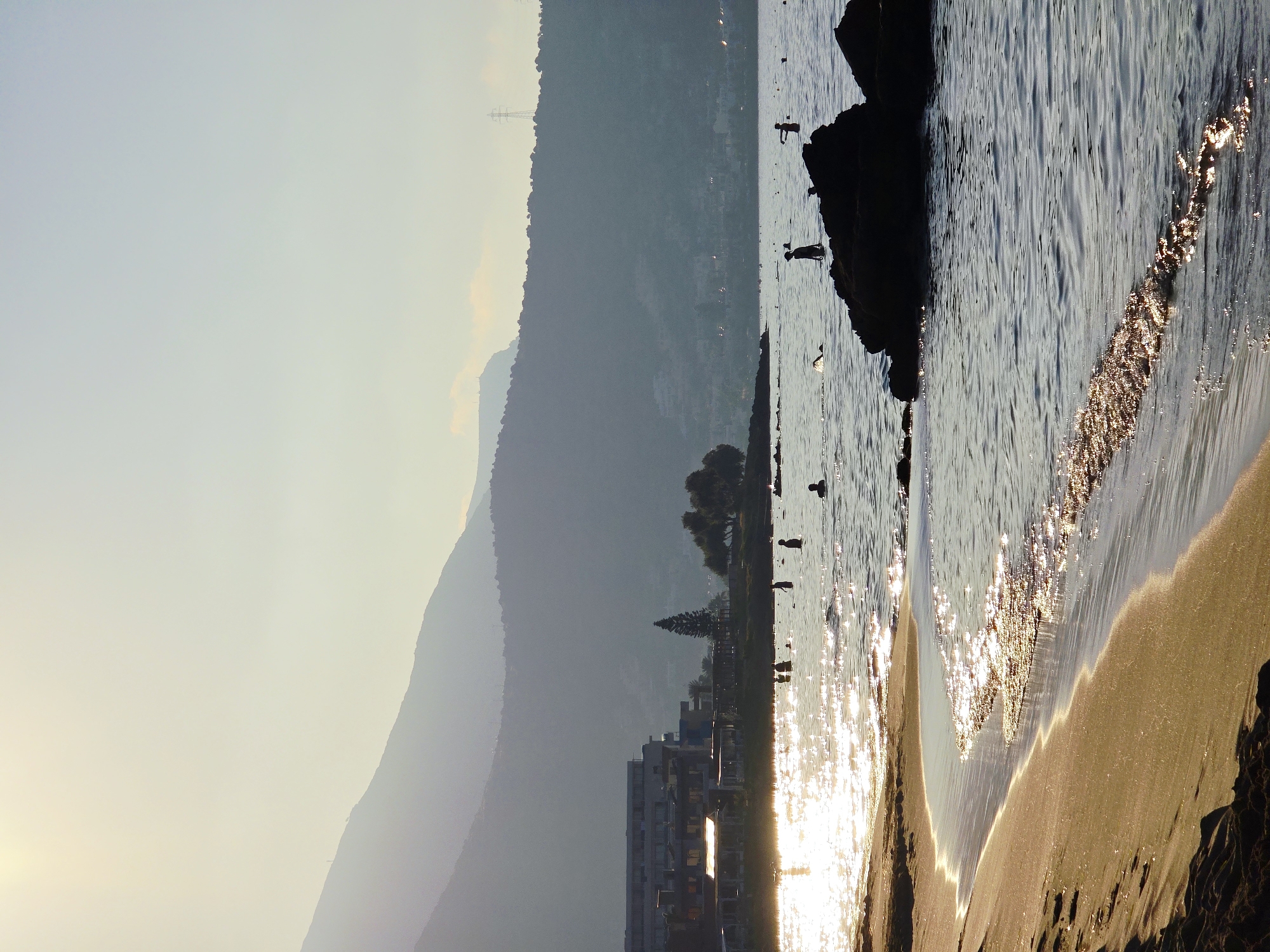 This is a photograph of a beach in Malia Municipal Unit, Greece, taken at dusk. The sun is setting, casting a warm glow on the water and sand. In the foreground, the gentle waves lap at the shore, their surfaces reflecting the golden light. Further out, several people are visible in the water, some swimming and others standing. In the mid-ground, a line of buildings runs along the coast, with palm trees and other vegetation in front of them. In the background, rolling hills rise, their peaks silhouetted against the pale sky. The scene is tranquil and serene, conveying a sense of peace and relaxation.