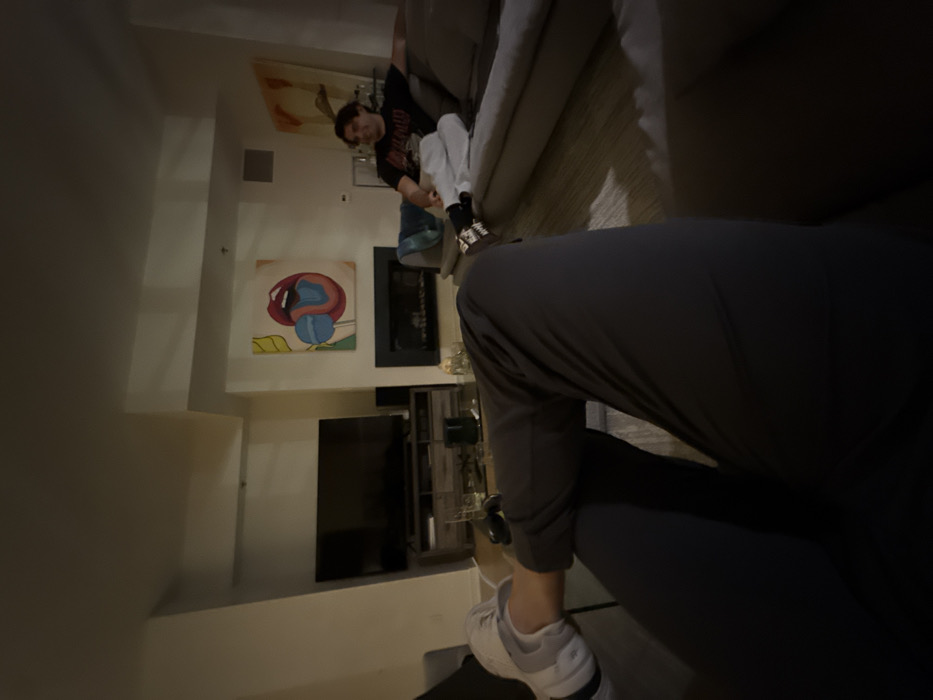 A man is on a sofa, partially visible, wearing a black t-shirt with a design. In the foreground, a person wearing gray athletic pants and white sneakers is seen. A television is mounted on the wall, beneath a painting featuring an abstract, colorful representation of lips and a sphere. A fireplace is situated between the artwork and the television. A cabinet with decorative items is placed below the television.