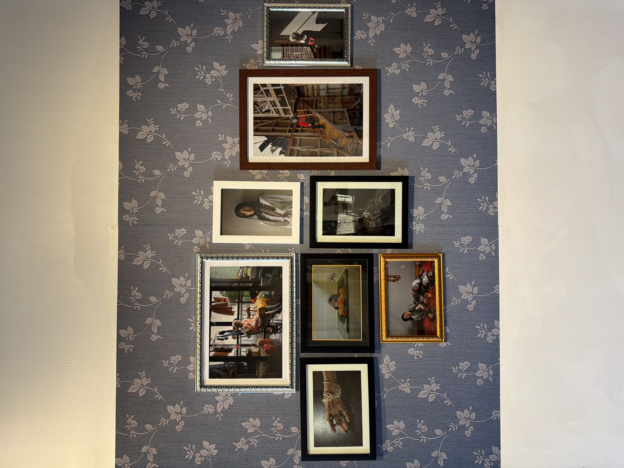 The image depicts a wall adorned with a floral wallpaper, upon which several framed photographs are displayed. The photographs are arranged in a cluster, creating a gallery-like presentation. The subjects of the photographs vary, including people in indoor and outdoor settings, as well as close-ups of hands and objects. One photograph shows a woman sitting in a rocking chair on a balcony with laundry drying in the background. Another features a woman wearing a headscarf. A third image shows a person on a staircase within a wooden structure. There are also close-ups of hands adorned with jewelry and a scene of a person in a bathtub. The lower portion of the wall is painted white. This display of framed photographs is likely located in a residential or commercial interior space, possibly a home, gallery, or cafe in Kediri, Indonesia, given the context of the request.