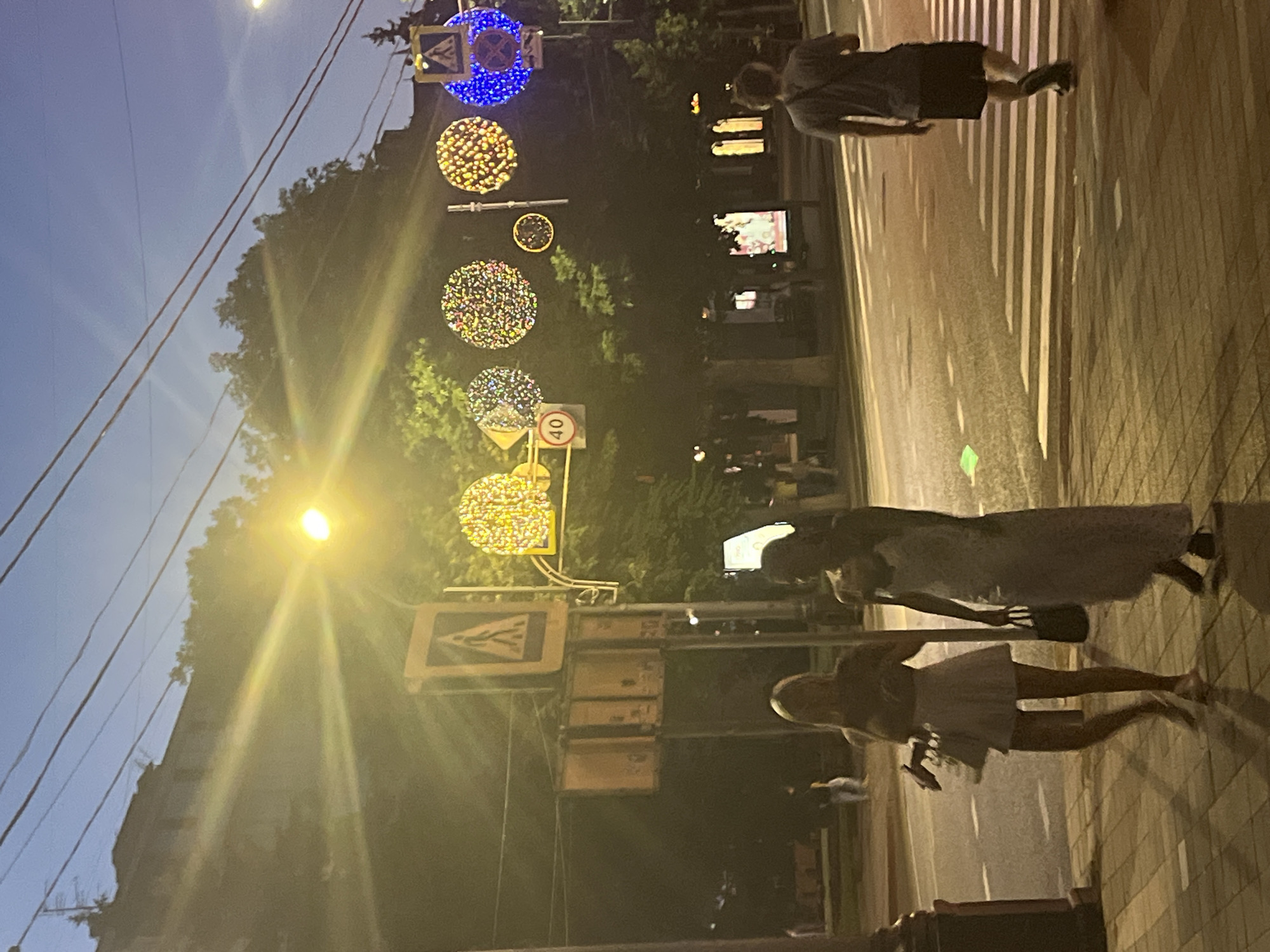In an urban setting at dusk in Krasnodar, Russia, several people are captured in motion. Two women, one in a light-colored dress and the other in a short white dress, are walking across a crosswalk. A man in a t-shirt and shorts is also visible, presumably on the same street. The scene is illuminated by streetlights and decorative circular light installations, suggesting a festive atmosphere, possibly a holiday or event. The sky is a deep blue, indicating the transition from day to night. Traffic signs are present, including a speed limit sign indicating "40" and a pedestrian crossing sign. The lighting creates a warm glow across the pavement and the surrounding trees. The image is oriented sideways, adding a unique perspective to the scene.