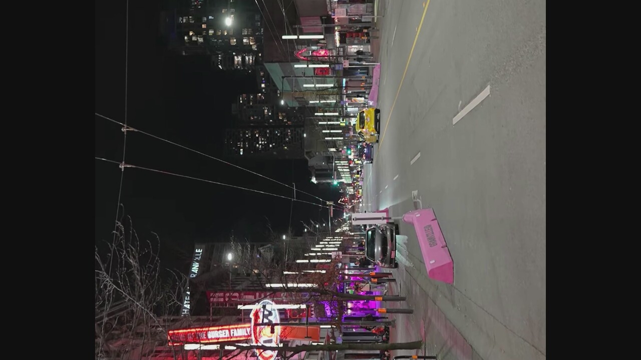 This is a night-time, eye-level shot of Granville Street in Vancouver, Canada. The street is lined with businesses displaying neon lights and vertical light poles. A few cars are visible on the road, including a yellow taxi and a dark-colored sedan. Pink traffic barriers are placed along the sidewalk. Pedestrians are walking on the sidewalk. The sky is dark, and the city lights from buildings in the background are illuminated. The overall scene depicts a lively urban environment at night.