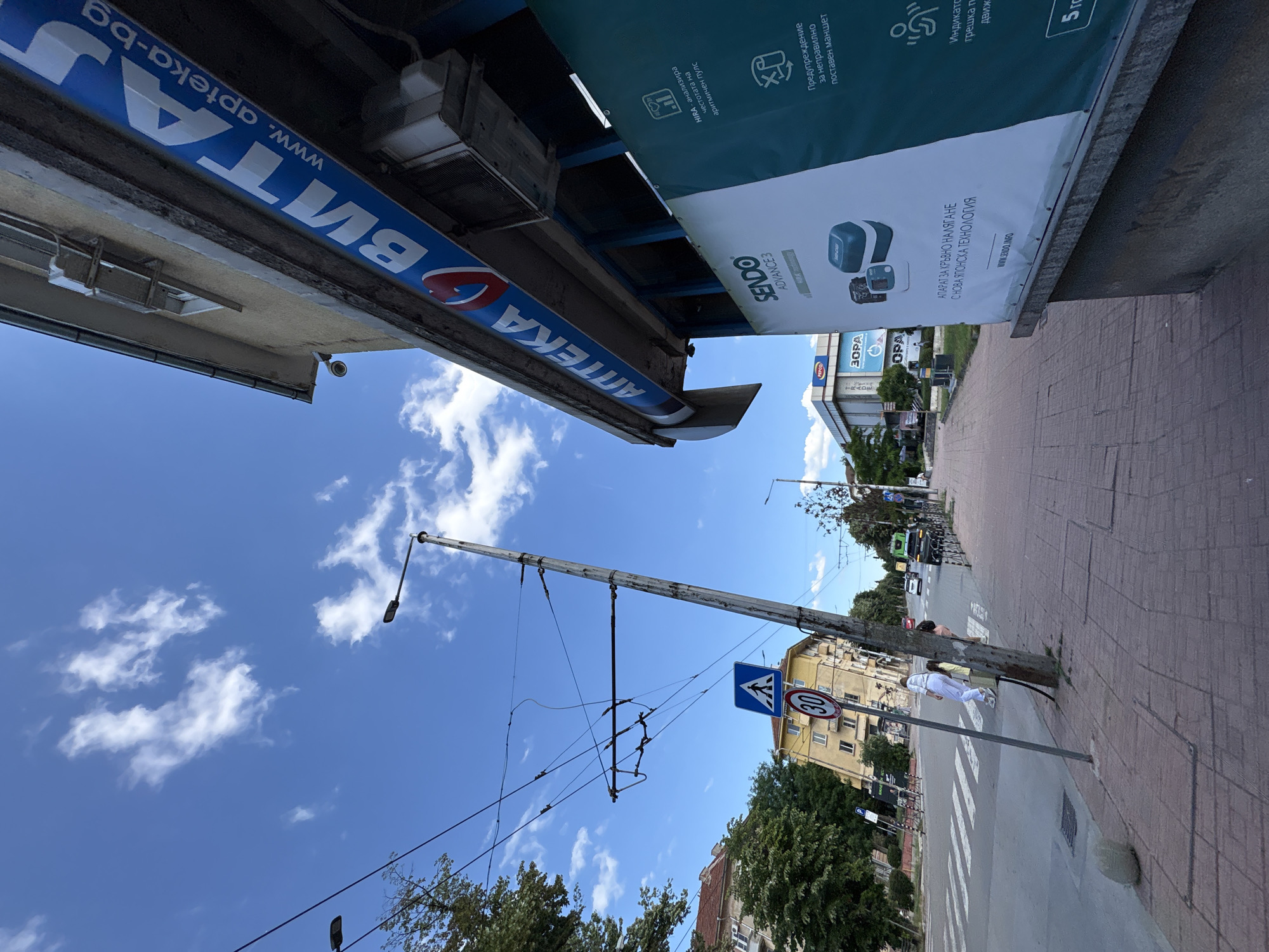 The image depicts a street scene in Vratsa, Bulgaria, featuring a pedestrian crossing, a tram line, and a pharmacy with a prominent blue and white sign. The sky is blue with scattered clouds.
In the foreground, a pedestrian crossing is visible on a paved road. A lamppost with electrical wires for a tram is positioned to the right of the crossing, leaning slightly. Two traffic signs are attached to the lamppost: a blue square sign with a white pedestrian symbol, and a circular sign with a red border and the number "30" in black, indicating a speed limit.
A person in white clothing and another person in yellow clothing are crossing the street from left to right. A woman with a yellow bag is walking on the sidewalk on the right side of the image.
To the right, the facade of a building is visible, featuring a blue banner with white lettering that reads "АПТЕКА ВИТА" and "www.apteka-bg". Below this banner is a large white poster advertising "Sendo Advance 3" blood pressure monitors, with text in Bulgarian describing its features, such as "HIRA - analysis of irregular pulse" and "Apparatus for blood pressure with new Japanese technology". The poster also shows images of the Sendo device.
In the background, other buildings, trees, and vehicles are present, including a green truck on the road. The environment appears to be an urban setting on a sunny day.