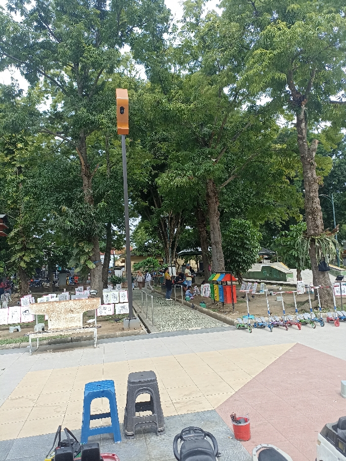 The image depicts a bustling outdoor public space, likely a park or town square in Tulungagung, Indonesia, under an overcast sky during the daytime. Large, mature trees with dense green foliage create a lush backdrop, providing shade throughout the area.
In the immediate foreground, the ground is paved with large tiles in shades of tan, grey, and reddish-brown. Two plastic stools, one blue and one black, are centrally positioned. Parts of what appear to be motorbikes or scooters are visible at the very bottom edges of the frame.
A tall, dark pole stands prominently, topped with an orange rectangular unit that resembles a sensor or camera. To the left of the pole, a white concrete bench is surrounded by numerous white display boards, each holding what appears to be artwork or drawings, possibly from a local exhibition or children's activity. People are scattered across the scene, some strolling along paved paths, others gathered in groups or sitting under the trees.
To the right of the central path, a line of colorful kick scooters is neatly parked, suggesting they are available for rent or use. Adjacent to these are several multi-colored bins (red, blue, yellow, green) with some indistinct labels. A distinct pathway made of small, rounded stones, characteristic of a reflexology walk, runs parallel to the scooter line, equipped with metal handrails. Further into the background, more individuals are seen enjoying the park amenities, with small structures and additional seating visible beneath the trees. The overall mood is relaxed, indicating a leisure or community gathering. No clearly legible text is visible, apart from the general impression of labels and artwork.
