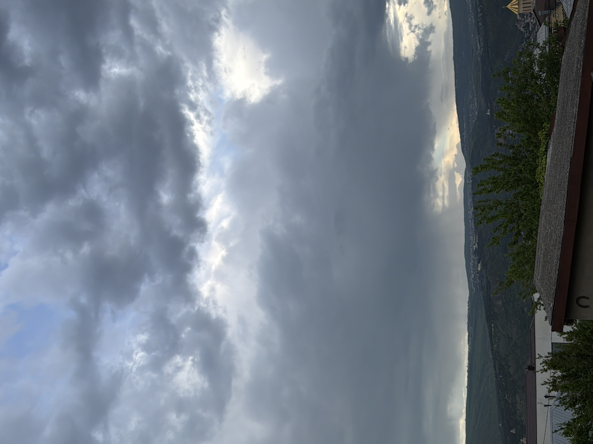 The image shows a landscape view from Tbilisi, Georgia, likely taken from an elevated position, possibly a balcony or rooftop. The sky is dominated by heavy, dark clouds, suggesting an overcast or stormy weather condition. Patches of lighter, brighter sky are visible, peeking through the cloud cover. A mountain ridge is partially visible in the midground, with green vegetation covering its slopes. A small portion of the city is visible along the ridge, including some buildings and possibly a golden dome. The foreground includes a portion of a building or structure, with a roof and some vegetation. The overall impression is one of a city nestled in a valley, under a dramatic and potentially threatening sky.