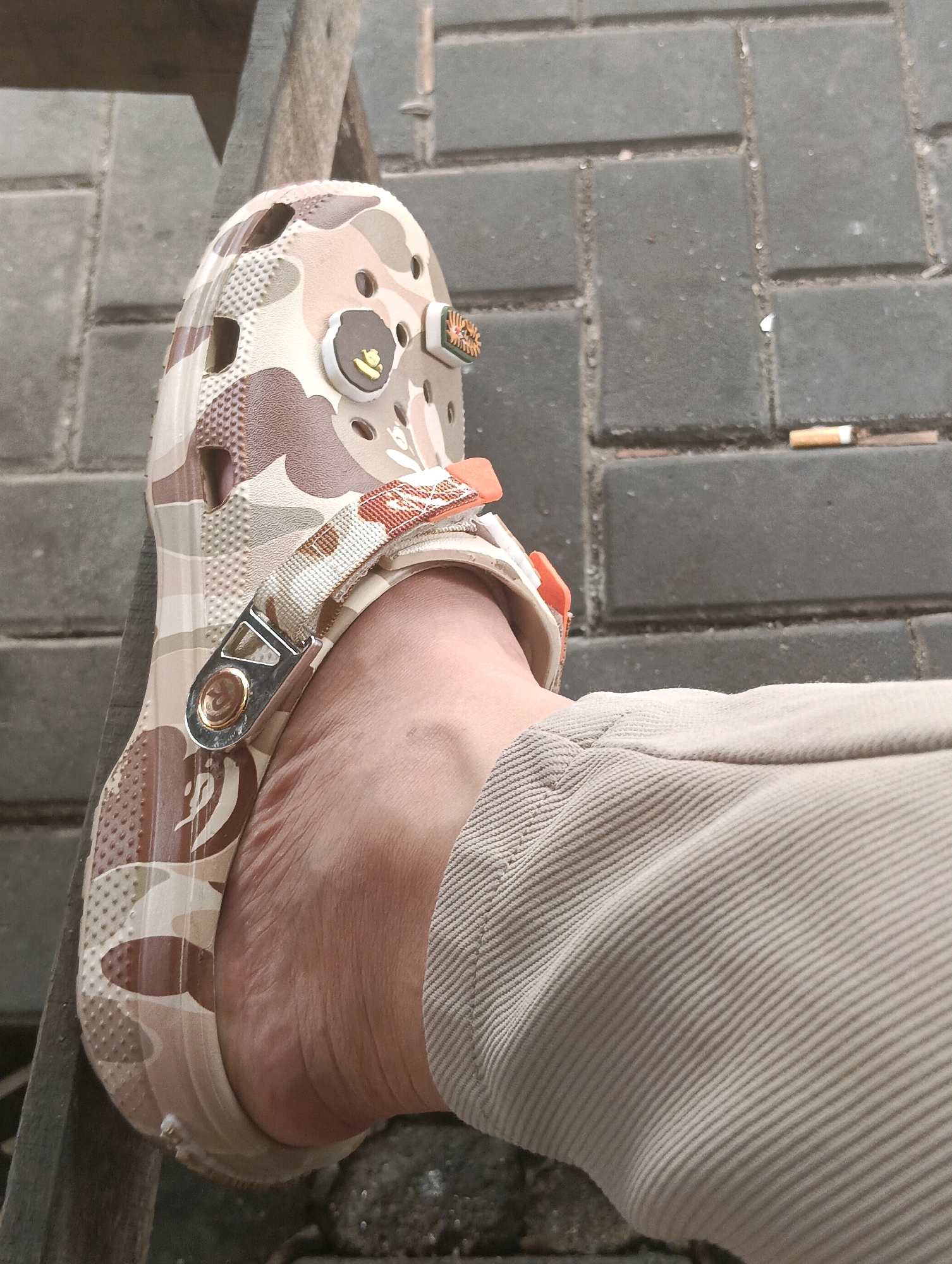 A close-up shot focuses on a person's left foot wearing a camouflage patterned Croc. The shoe has orange accents and a small, brown and gold charm depicting a face. The person is wearing tan corduroy pants. The foot is resting on a wooden surface, and the background consists of gray brick paving stones with a discarded cigarette butt on the ground. The scene appears to be outdoors, possibly in a public area within Kuching, Malaysia.