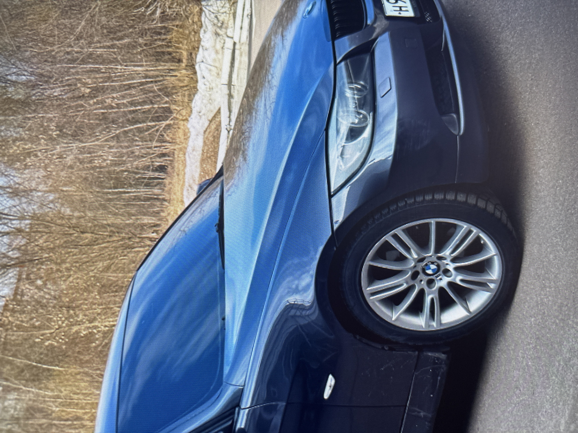 A dark blue BMW sedan is parked on an asphalt road. The car has a dent on the front passenger side fender. The background shows a line of bare trees and some patchy snow on the ground, suggesting a cold-weather environment. The vehicle's front right wheel and part of the front bumper are visible. The license plate is partially obscured but shows Cyrillic characters. The scene appears to be outdoors, possibly in a suburban or park area, given the natural elements in the background. No people are present in the image. The lighting suggests it might be daytime.