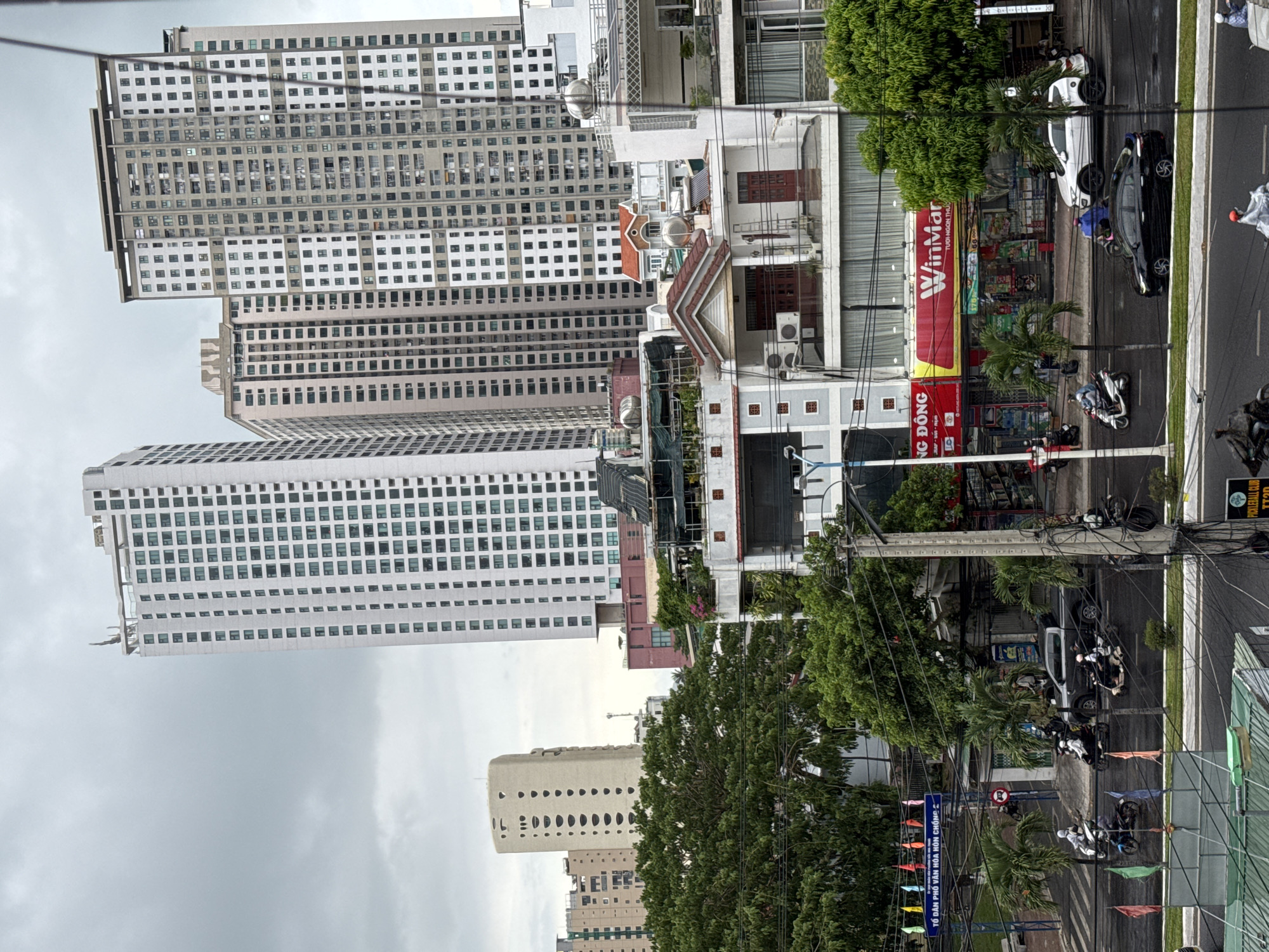 The image shows a street scene in Nha Trang Ward, Vietnam, dominated by tall buildings. The sky is overcast. Several high-rise buildings are visible, characterized by numerous windows and a modern architectural style. A WinMart store stands out with its red and white sign. Cars and motorbikes are parked or moving along the street. Utility poles and wires crisscross the scene. Signs indicating local cultural and community information are present, including one that reads "TỔ DÂN PHỐ VĂN HÓA HÒN CHỐNG" and "NG ĐÔNG". Small flags are displayed along the street. Green trees are interspersed between the buildings and along the roadside. A pickleball club is also listed.
