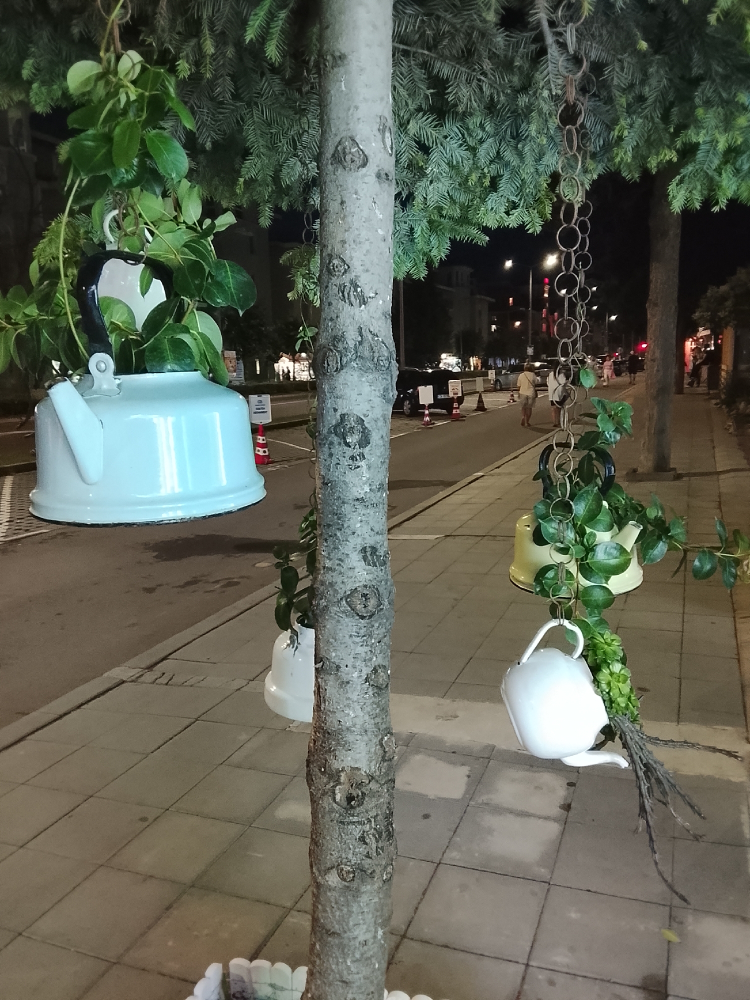 In Nesebar, Bulgaria, a tree trunk is adorned with hanging kettles serving as planters. The large, white enamel kettle on the left is filled with vibrant green foliage. To its right, a rusty metal chain hangs, supporting a yellow kettle also containing plants. Below the yellow kettle, a small white pitcher, containing green succulents and dried branches, is suspended. The scene is set at night, with streetlights illuminating a paved sidewalk and a road in the background where a few pedestrians are visible.