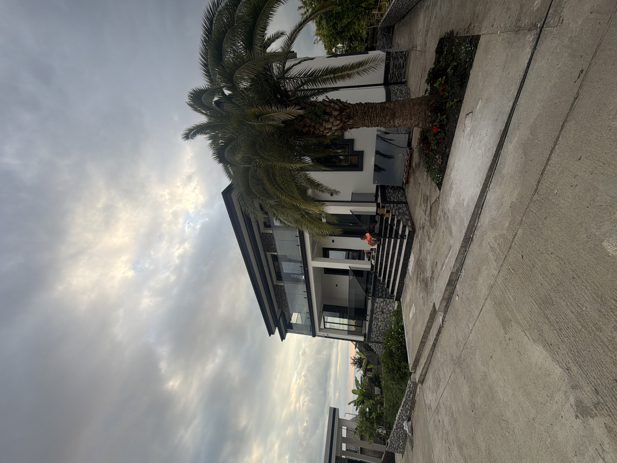 An exterior, wide-angle, eye-level shot captures a modern, two-story white house with dark trim and glass balconies. The house is situated on a slight incline, with a concrete pathway leading up to a set of dark-tiled stairs. A large palm tree stands prominently in front of the house, its fronds reaching towards the overcast sky. To the left of the house, another modern structure with white columns and a flat roof is partially visible. Lush green vegetation and some flowers are planted around the base of the palm tree and in front of the structures. A person wearing an orange shirt is seated on the stairs. The sky is filled with dramatic, textured clouds. The scene appears to be in a residential area, possibly near a coast given the presence of palm trees and the distant, hazy view of water. The overall impression is one of a newly constructed or renovated property.