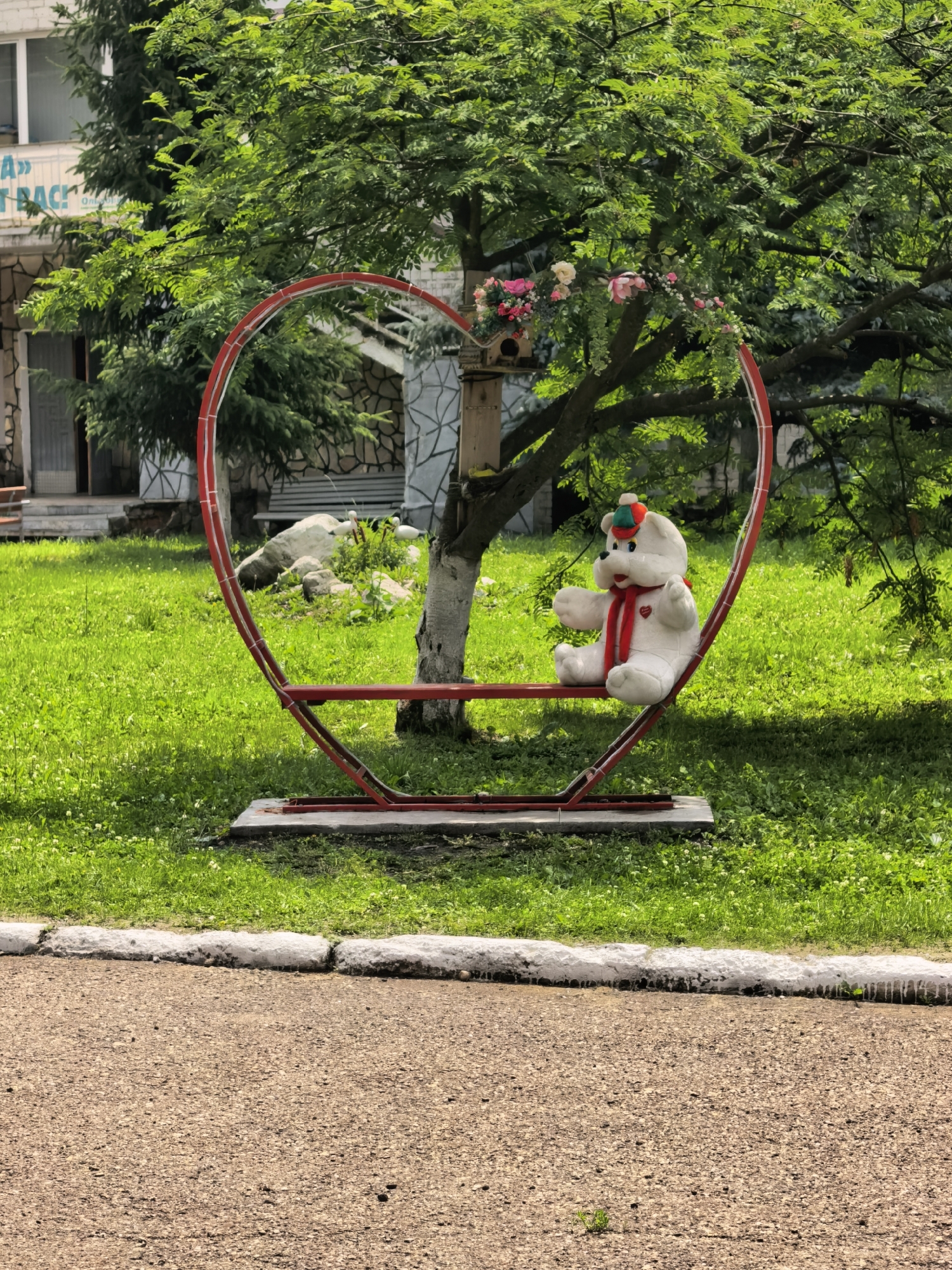 In Sterlitamak, Russia, a white teddy bear wearing a red scarf and a multicolored hat sits on a red heart-shaped swing in a grassy area near a building. The swing is supported by a metal frame. There are trees with green leaves and a birdhouse hanging from a branch. The ground in front of the swing is covered in green grass, and a paved path is visible in the foreground. The building in the background appears to be made of stone and has windows.