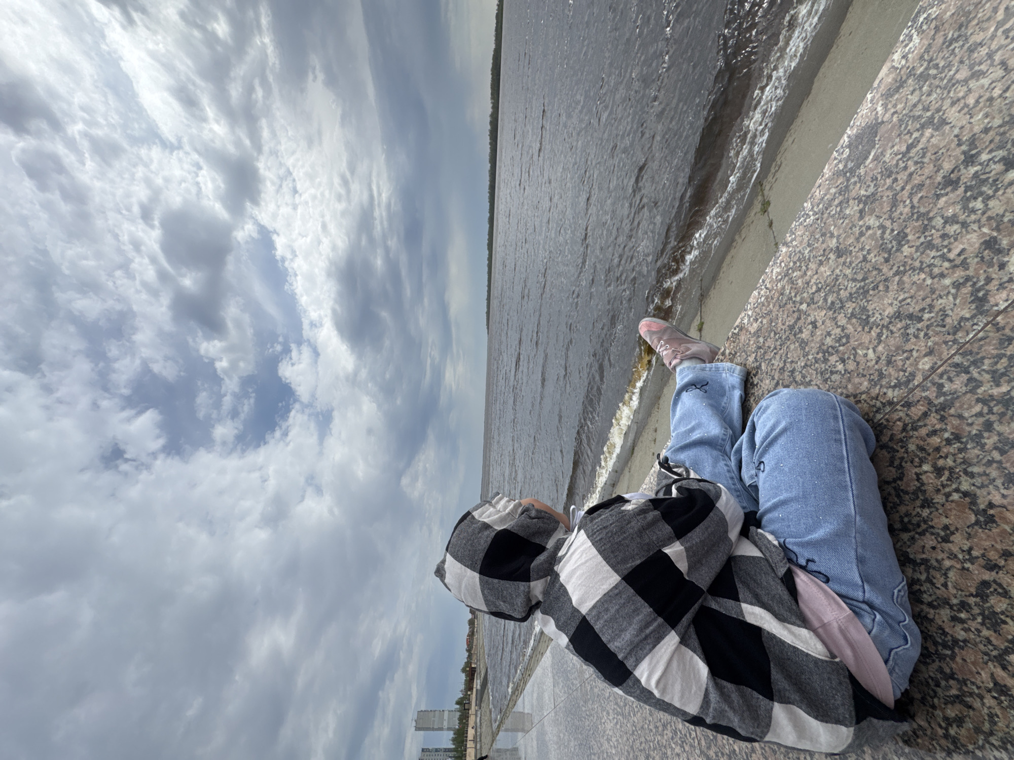 A person wearing blue jeans, pink sneakers, and a black and white checkered hooded shirt sits on a granite barrier next to a body of water under a cloudy sky. The person faces the water. The photo appears to be taken near a riverbank in Nizhnevartovsk, Russia, based on the skyline visible in the distance. Waves from the water lap against the concrete barrier.