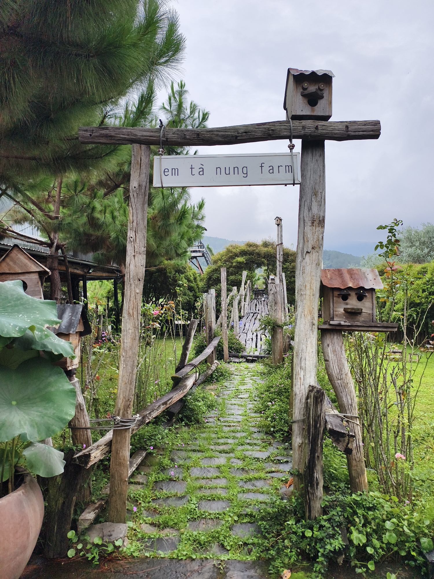 The image shows the entrance to "Em Ta Nung Farm" in Thôn 5, Vietnam. The entrance is marked by a rustic wooden archway with a sign bearing the farm's name. Atop each vertical post of the archway is a wooden birdhouse resembling a face. A stone path, partially covered in greenery, leads into the farm. The path is flanked by a weathered wooden fence on either side. Lush vegetation surrounds the path, including flowering plants, shrubs, and large lotus leaves in a pot on the left. In the background, a wooden bridge-like structure can be seen, along with trees and a cloudy sky.