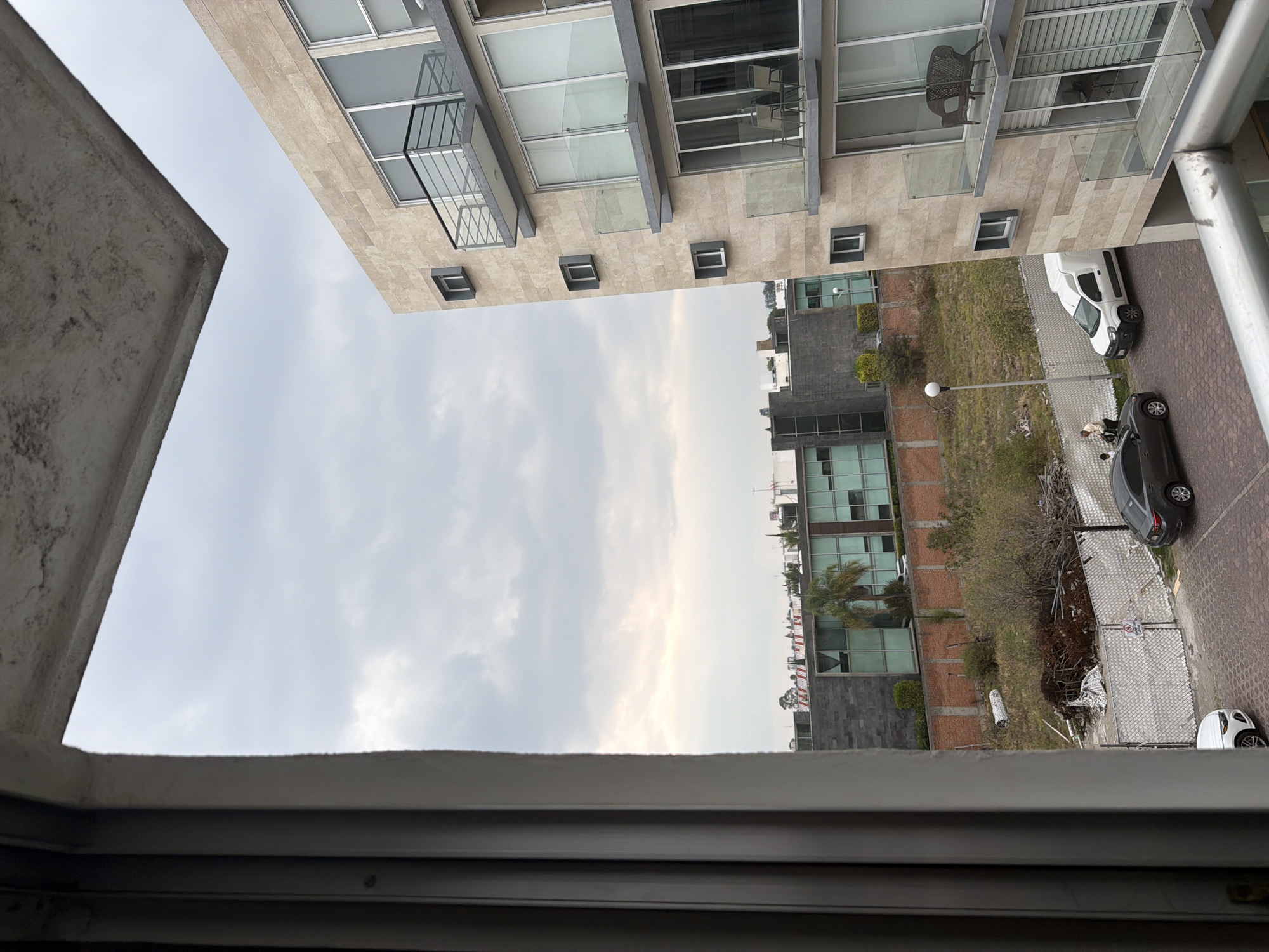 The image depicts an upward view from a window, likely in San Andrés Cholula, Mexico, characterized by urban development and partly cloudy skies. Two multi-story buildings are visible, one with a modern facade featuring balconies, large windows, and neutral-toned stone cladding, and another with a mix of glass and darker materials. A landscaped area is present between the buildings, featuring grass, bushes, and a few trees, and a chain-link fence. A street with parked cars is to the side. The foreground features the frame of the window, which is made of metal. No people are clearly identifiable.