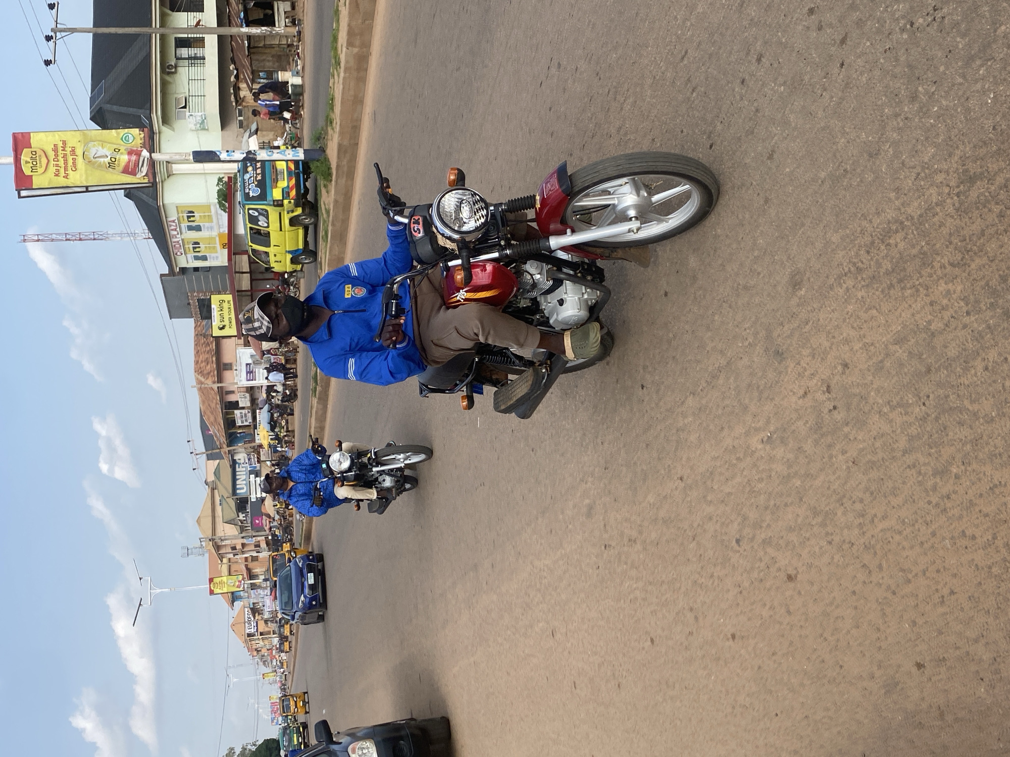 The image depicts a street scene in Sabon Tasha, Nigeria, during daylight. A man wearing a blue jacket, brown pants, and a black face mask is riding a red motorcycle with silver accents. He is looking forward, focused on the road ahead. Another motorcyclist, also wearing a blue jacket, is visible further down the road, to the left of the main subject. The street is paved and shows signs of wear, with a reddish-brown hue. Several vehicles, including cars and a yellow van, are present on the road. Buildings line the right side of the street, with shops and signage indicating commercial activity. The sky is partly cloudy. The overall atmosphere suggests a bustling urban environment with ongoing traffic.