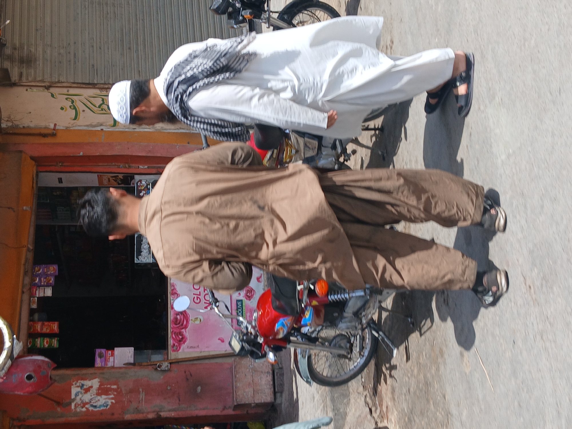 Two men stand next to motorcycles on a street in Quetta City Tehsil, Pakistan. One man wears a white thobe with a white prayer cap and a black and white patterned scarf. The other man wears a brown salwar kameez. Both men wear sandals. The motorcycles are parked next to a shop with a red and faded facade. The sidewalk is made of concrete.