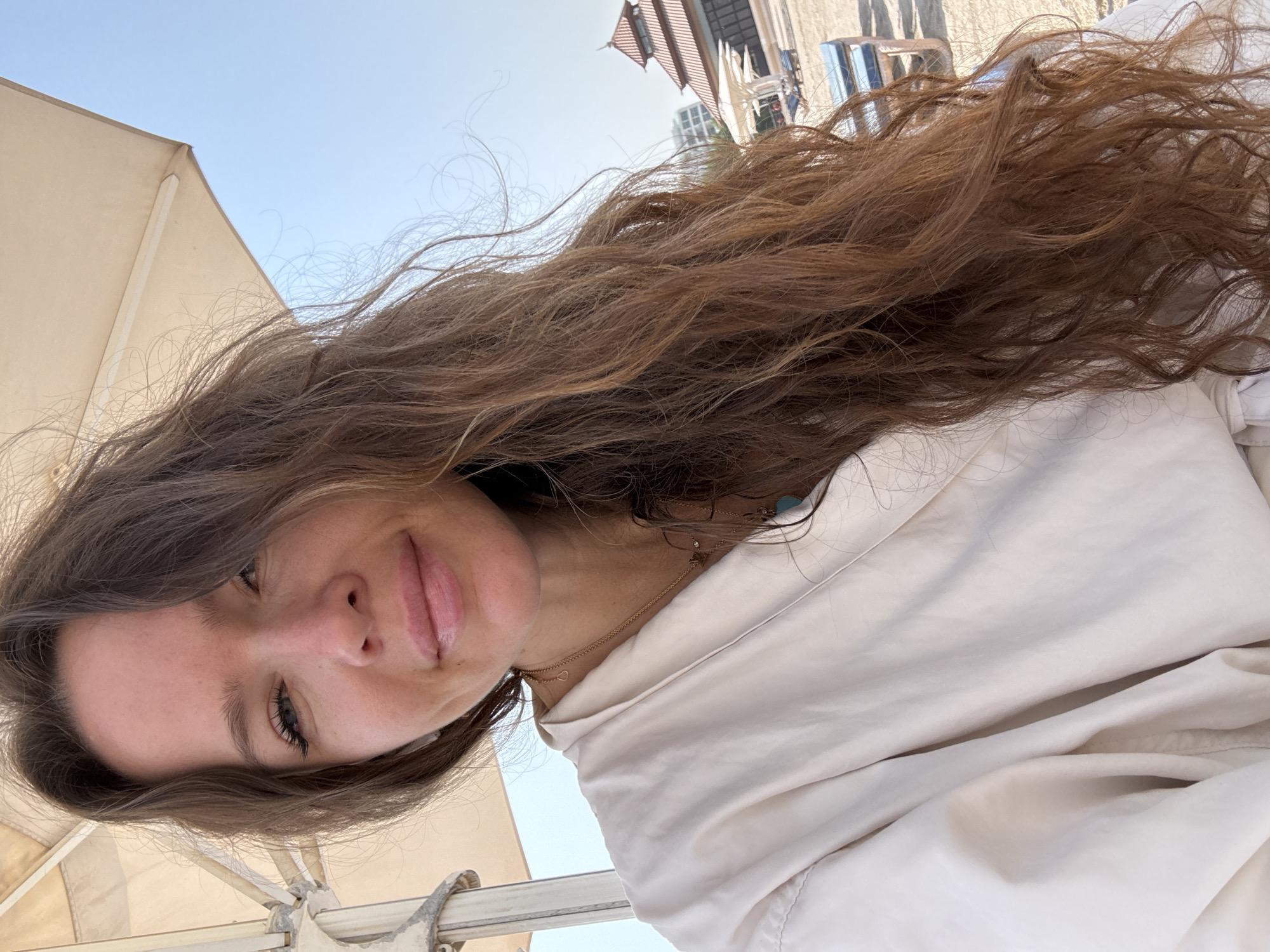 A medium shot captures a woman taking a selfie outdoors on a sunny day in Dubai, United Arab Emirates. The woman, positioned in the foreground, is wearing a light-colored robe and has long, wavy brown hair that extends down her right side. She is looking at the camera with a slight smile. Her face is illuminated by natural sunlight, and her makeup is subtle. She is wearing delicate gold necklaces.
Behind her, an umbrella provides shade, and beyond that, a sandy area with lounge chairs and a building with a reddish-brown tiled roof is visible. The sky is clear and blue, indicating fair weather. The background suggests a beach or resort setting within Dubai. The overall scene is casual and relaxed.
