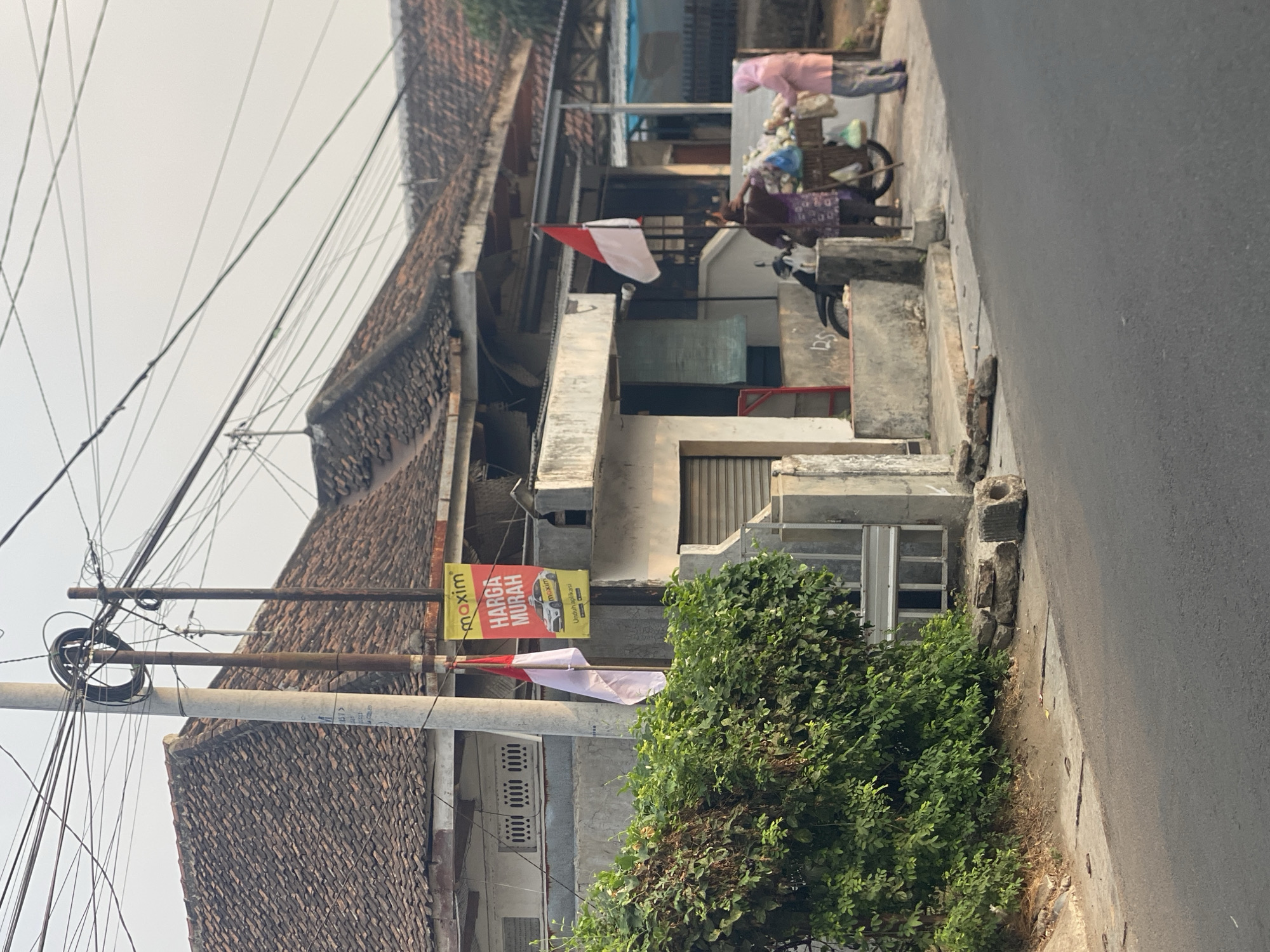 The image depicts a street scene in Kediri City, Indonesia, likely in a residential area. In the foreground, a paved road is visible. On the left side of the road, a dense green bush grows alongside a concrete structure. A utility pole stands near the bush, with numerous electrical wires crisscrossing overhead. A small Indonesian flag, white and red, is attached to the pole. A yellow banner advertising "Maxim" and "HARGA MURAH" (cheap prices) with a car image is also displayed on the pole.
Behind the bush and pole, a single-story house with a tiled roof is partially visible. A concrete wall with an opening and a metal gate runs along the front of the property. Further to the right, two individuals are seen near a motorized cart filled with goods. One person, wearing a purple patterned outfit, appears to be interacting with the cart. The other person, dressed in a pink headscarf and pink top, stands beside the cart. A motorcycle is parked behind the cart. The overall environment suggests a warm, possibly early morning or late afternoon, atmosphere given the lighting.