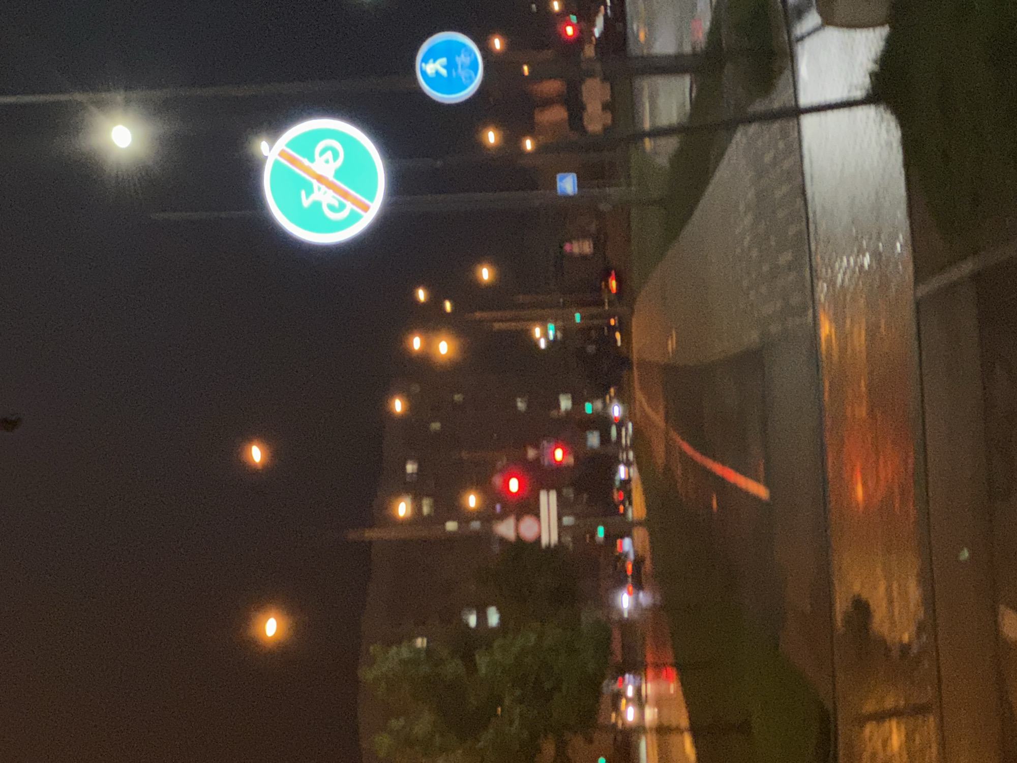 The image, taken at night in Бараўлянскі сельскі Савет, Belarus, shows a street scene. In the foreground, there's a pedestrian path beside a reflective glass wall. In the middle ground, two traffic signs are visible: one indicating "No Bicycles Allowed" and another showing a pedestrian and bicycle path. In the background, there's a multi-story building with illuminated windows, streetlights, and traffic lights. The overall atmosphere is dark with artificial lighting. There are no people visible.