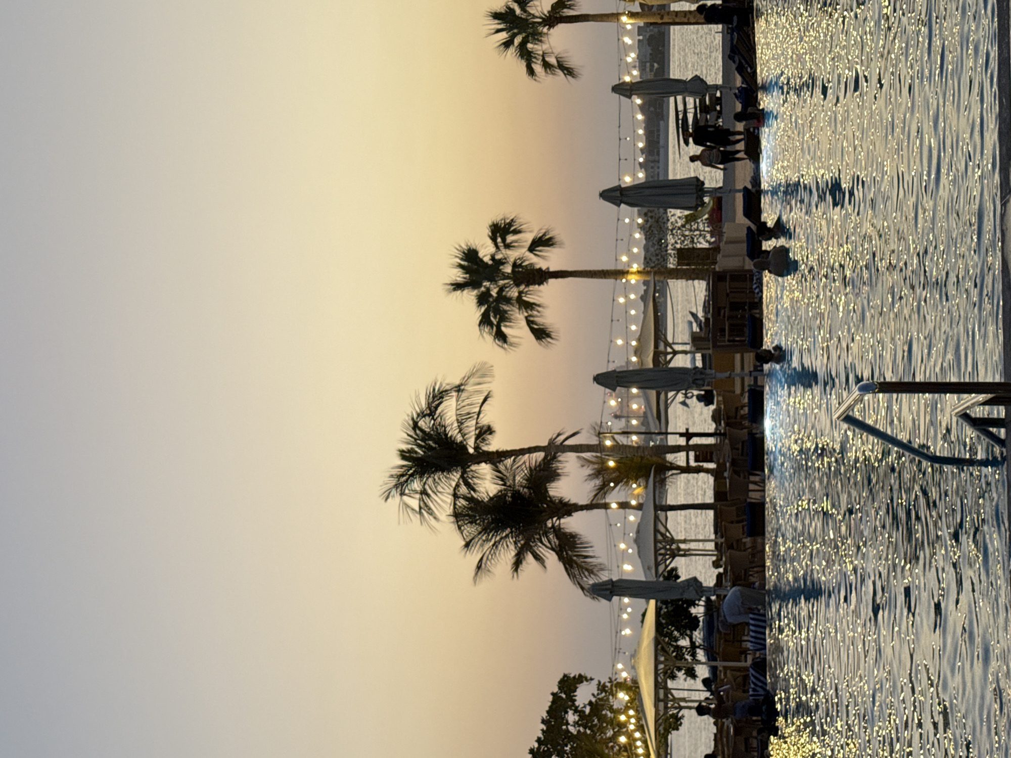 A scenic view of a swimming pool at dusk, likely in Dubai, United Arab Emirates, with palm trees and a beachside restaurant in the background. The sky is a gradient of soft orange and yellow, indicating sunset or sunrise. The pool water is rippled, reflecting the warm evening lights. Several people are visible in and around the pool, some swimming, while others are seated at tables or standing near the shore. The background features a row of palm trees, beach umbrellas, and string lights, suggesting a resort or a popular leisure area. Further in the distance, a body of water, possibly the sea, is visible, with buildings and structures lining the horizon. The overall ambiance is relaxed and tropical, with a touch of urban sophistication characteristic of Dubai.