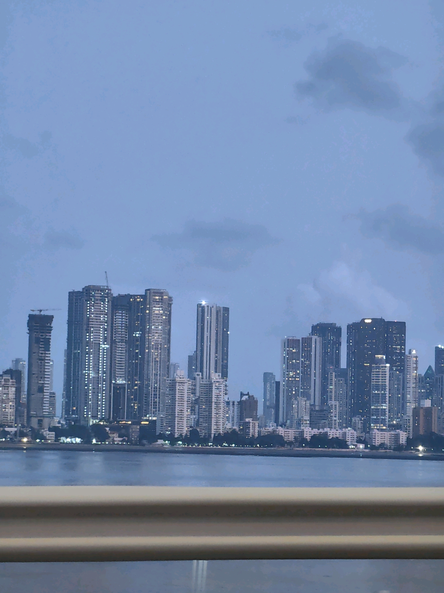 The image depicts the Bandra-Worli Sea Link in Mumbai, India, at dusk. A vast expanse of calm water, likely the Arabian Sea, fills the lower half of the frame, reflecting the dim evening light. In the background, the Mumbai skyline is illuminated by the lights of numerous skyscrapers. The sky is a muted blue, with subtle grey clouds creating a diffused light. A beige-colored railing runs horizontally across the foreground, suggesting the viewpoint is from a bridge or elevated walkway. The overall scene conveys a serene and urban atmosphere.