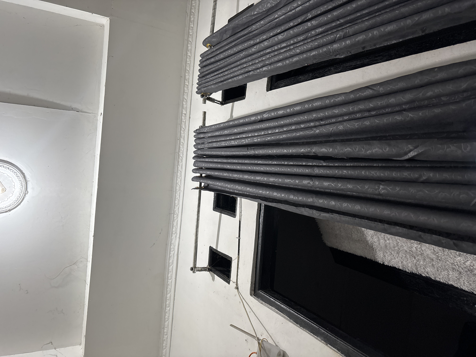 This is a photograph taken from a low angle looking up at a ceiling and the upper portion of a wall in what appears to be an indoor residential setting. The ceiling is white and shows some signs of wear, including minor cracks and discoloration. A decorative ceiling fixture is visible at the top center of the image. The wall below the ceiling is also white and features ornate white molding along the top edge.
Dominating the middle of the image are two sets of dark grey curtains, hung from metal rods. The curtains have a subtle embossed pattern. To the left of the curtains, the wall has two small, dark rectangular openings, possibly vents or small windows. A metal rod is mounted above these openings, and a light-colored cord extends from the left side of the frame towards the wall.
Below the openings and partially obscured by the curtains is a doorway with a black frame. Inside this doorway, a light-colored, plush rug is visible. The overall impression is of a room that may be undergoing renovation or has some unfinished elements, given the visible cracks on the ceiling and the functional rather than decorative appearance of the window openings and curtain rods.
The image does not contain any people or specific indicators of its exact location within North Jakarta, Indonesia, beyond it being an interior space.