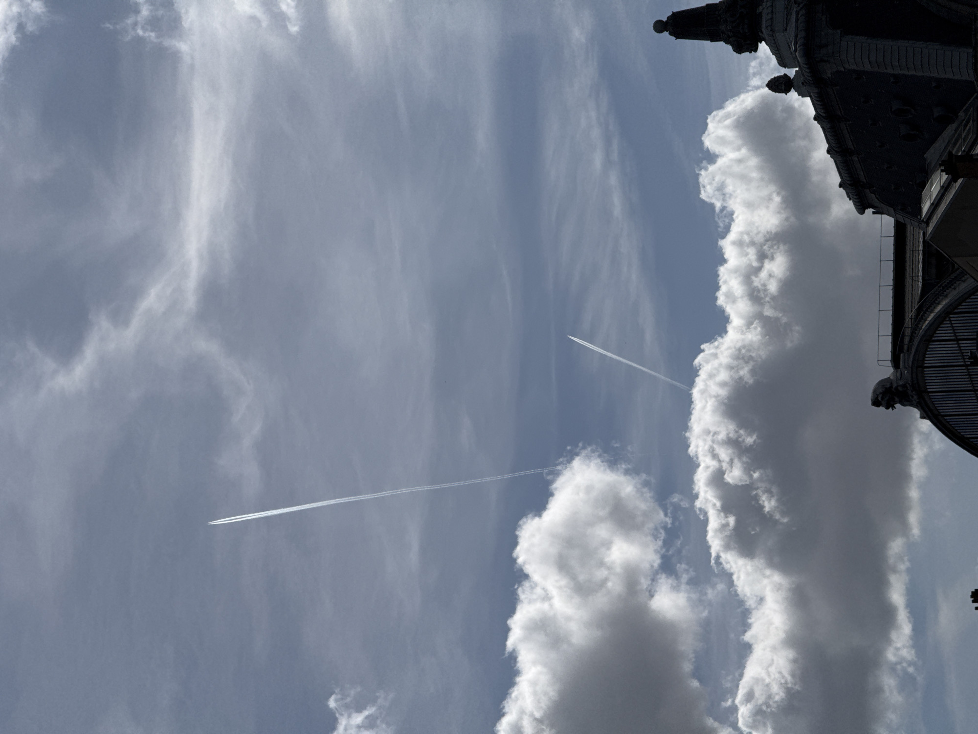The image displays a vibrant blue sky adorned with various cloud formations and aircraft contrails, with an ornate building partially visible on the right.
The sky is the dominant subject, featuring both dense, bright white and grey cumulus clouds, primarily forming a large vertical mass along the right side of the frame, and more delicate, wispy cirrus clouds spread across the lighter blue sections of the sky. Two distinct, long white contrails, left by high-flying aircraft, traverse the sky diagonally. One stretches from the lower-left quadrant towards the upper-right, while another, shorter contrail is situated higher up in the mid-left area, suggesting active air traffic.
On the far right side of the image, a dark, intricately detailed architectural structure is partially silhouetted against the bright sky. It appears to be part of a grand building, possibly featuring a dome or a tall spire with decorative elements, cornices, and balconies or railings. This classical architectural style is consistent with the provided location context of Paris, France.
The scene depicts a bright daytime setting, indicated by the clear blue sky and the illumination of the clouds. The weather appears fair with good visibility. No visible text is present in the image.
