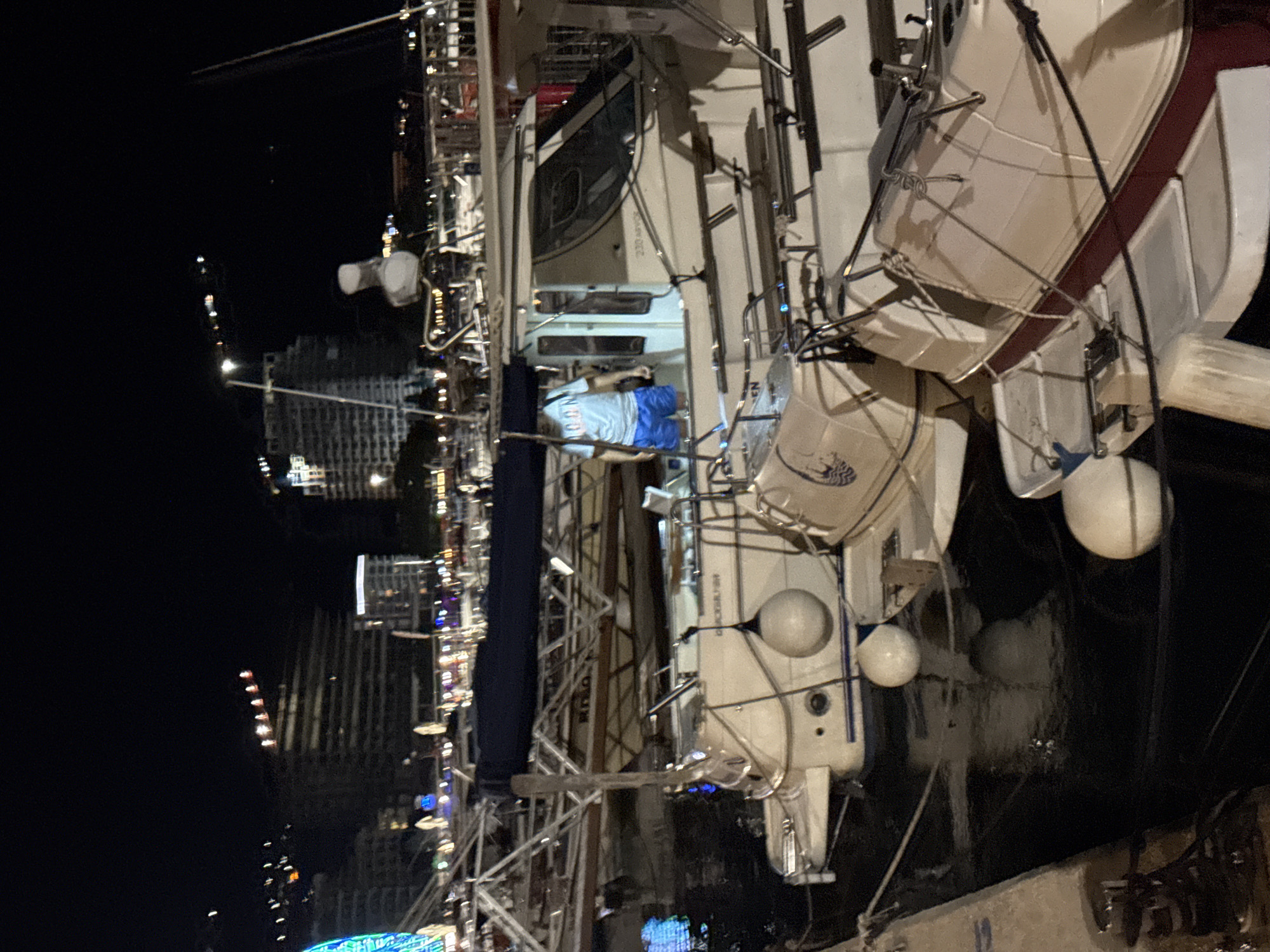 At night, a person wearing a light blue t-shirt and blue shorts stands on a white boat in a marina. The boat has a dark blue canopy and a small cabin. Mooring buoys are visible around the boat. In the background, the city of Budva, Montenegro, is illuminated with lights. Buildings and construction cranes are visible, along with a Ferris wheel on the left side of the image. The water is dark and reflects the lights of the city and the boats.