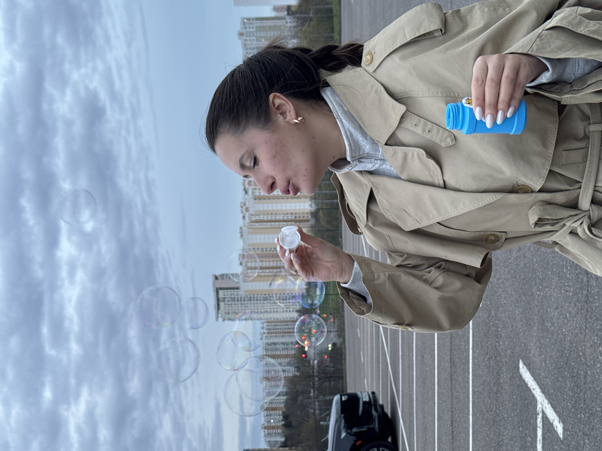 A woman with dark hair braided and styled in a bun, wearing a beige trench coat and a gray hoodie, is blowing bubbles outside. She is holding a small blue bottle in her right hand and a wand with bubble solution in her left. Several bubbles are floating in the air in front of her.
The location appears to be an asphalt parking lot, marked with white lines, with a car parked in the background. Behind the parking lot, a row of tall apartment buildings can be seen. The sky is overcast. Based on the architecture of the buildings and other visual cues, the location is likely within the city of Moscow, Russia.