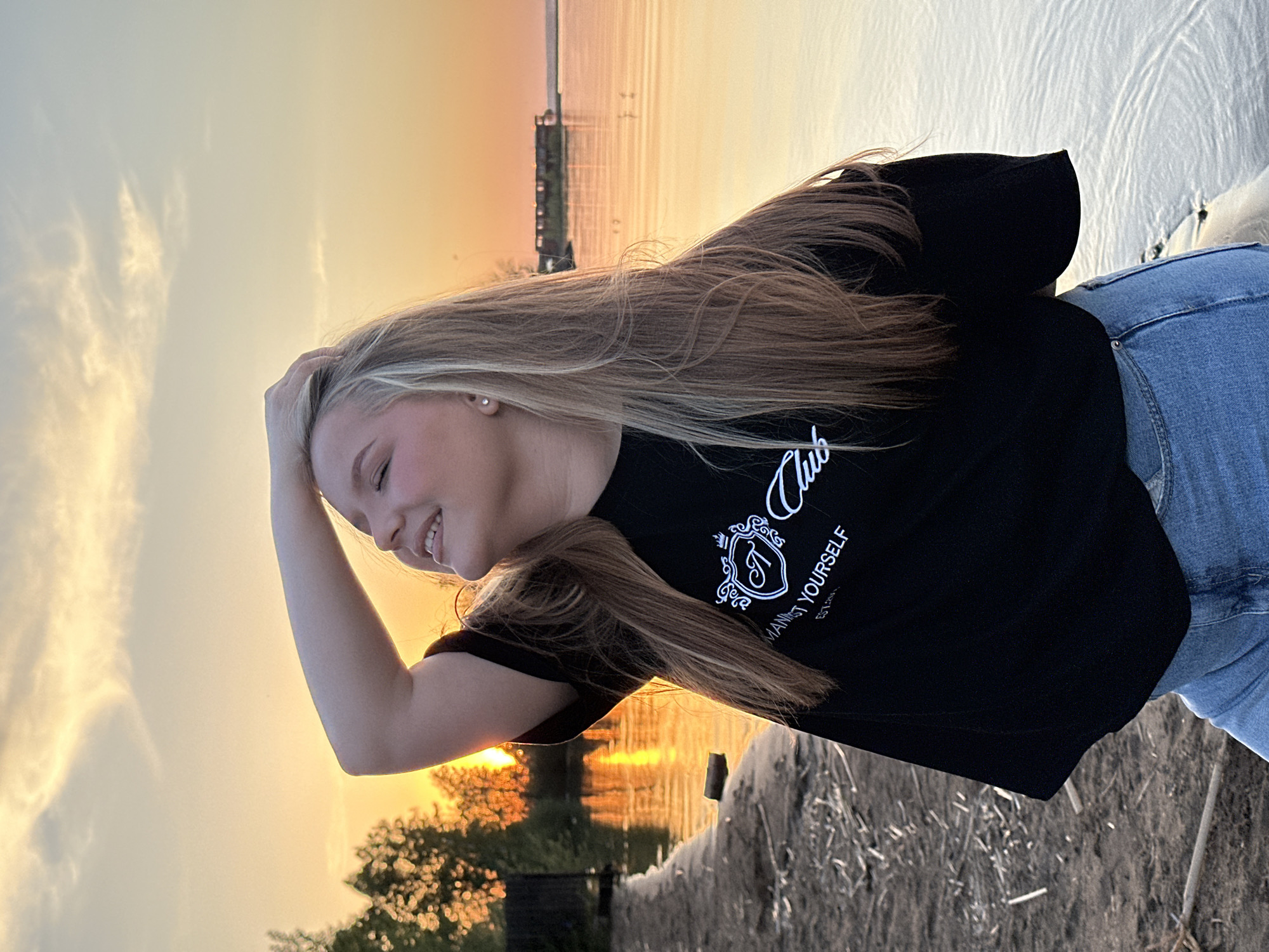 A young woman poses outdoors during sunset. She has long, blond hair and is wearing a black t-shirt with white lettering and a crest-like design. She is wearing blue jeans and has her left hand touching her hair. She is smiling slightly and looking to the right of the frame. In the background, there is water, possibly a river or lake, with golden reflections of the sunset. A shoreline with vegetation is also visible. The location might be Chaikovsky, Russia, based on the prompt. The environment is characterized by warm, sunset lighting.