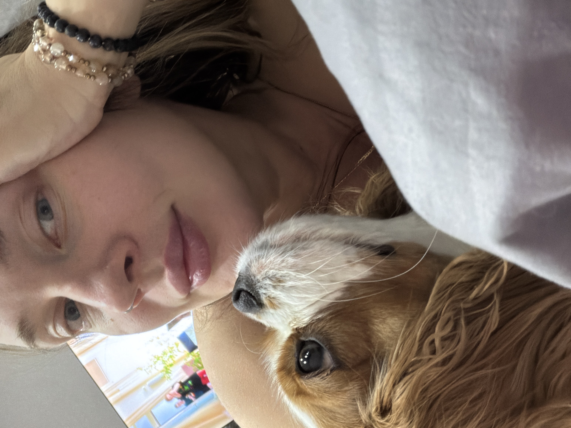 A woman is lying down next to a Cavalier King Charles Spaniel. The woman is fair-skinned with light-colored eyes. She is wearing bracelets on her left wrist and a necklace. The dog is mostly brown and white. The woman appears to be inside near a monitor showing light colors. The location is unknown and cannot be determined from the provided image.