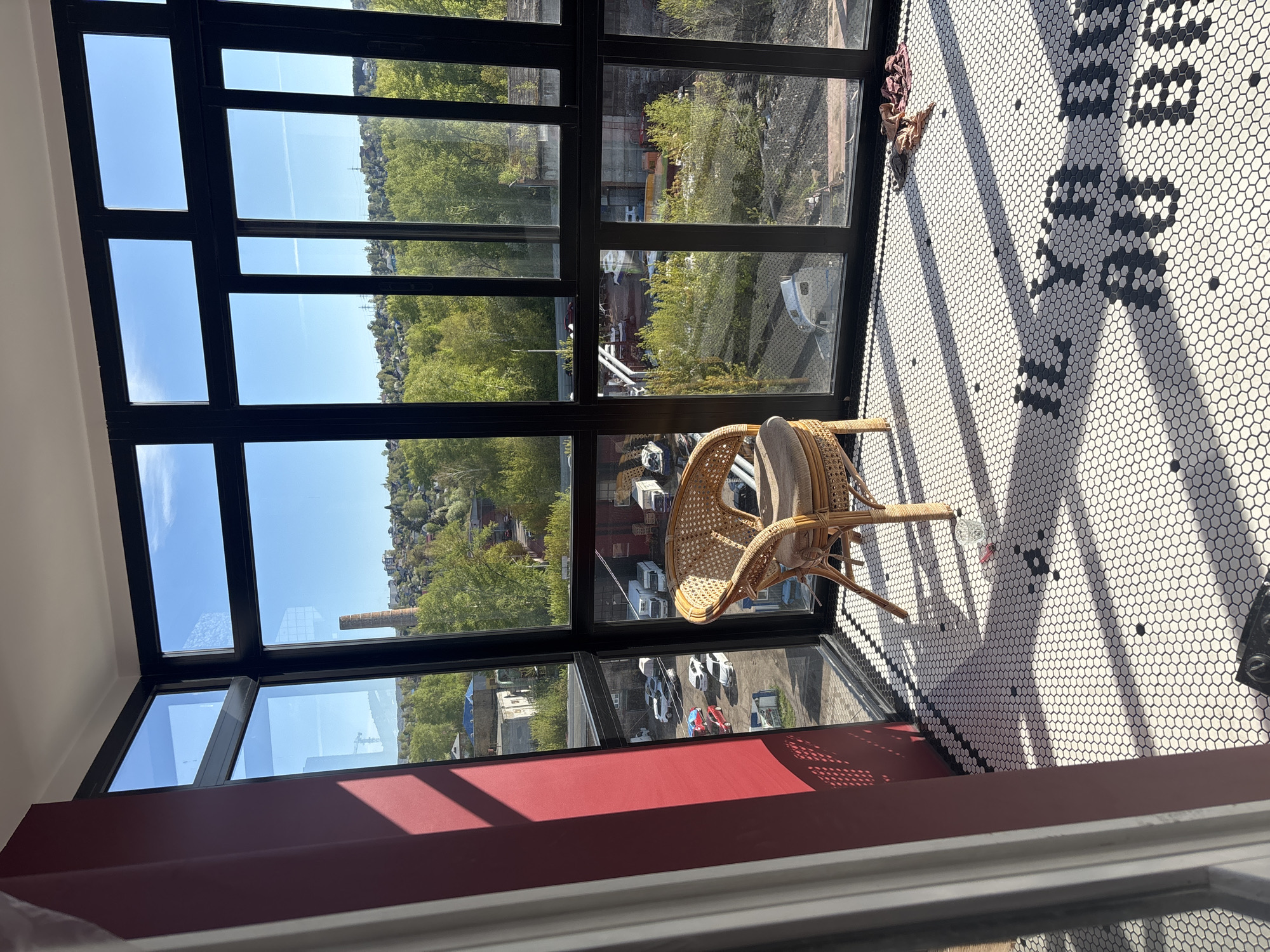 A low-angle, interior view depicts a sunlit space with a woven chair positioned near a large window offering a panoramic view of an urban landscape.
The chair, crafted from woven material, sits slightly angled towards the window. The floor is covered in a pattern of small, white hexagonal tiles, with black tiles forming a design.
The window is segmented by black frames, revealing a cityscape beyond. The view includes buildings, greenery, and roads.
Based on the industrial chimney visible and the general layout of the scene, the location may be Krasnoyarsk, Russia, specifically referencing the Museum Center "Plotmir" located there.